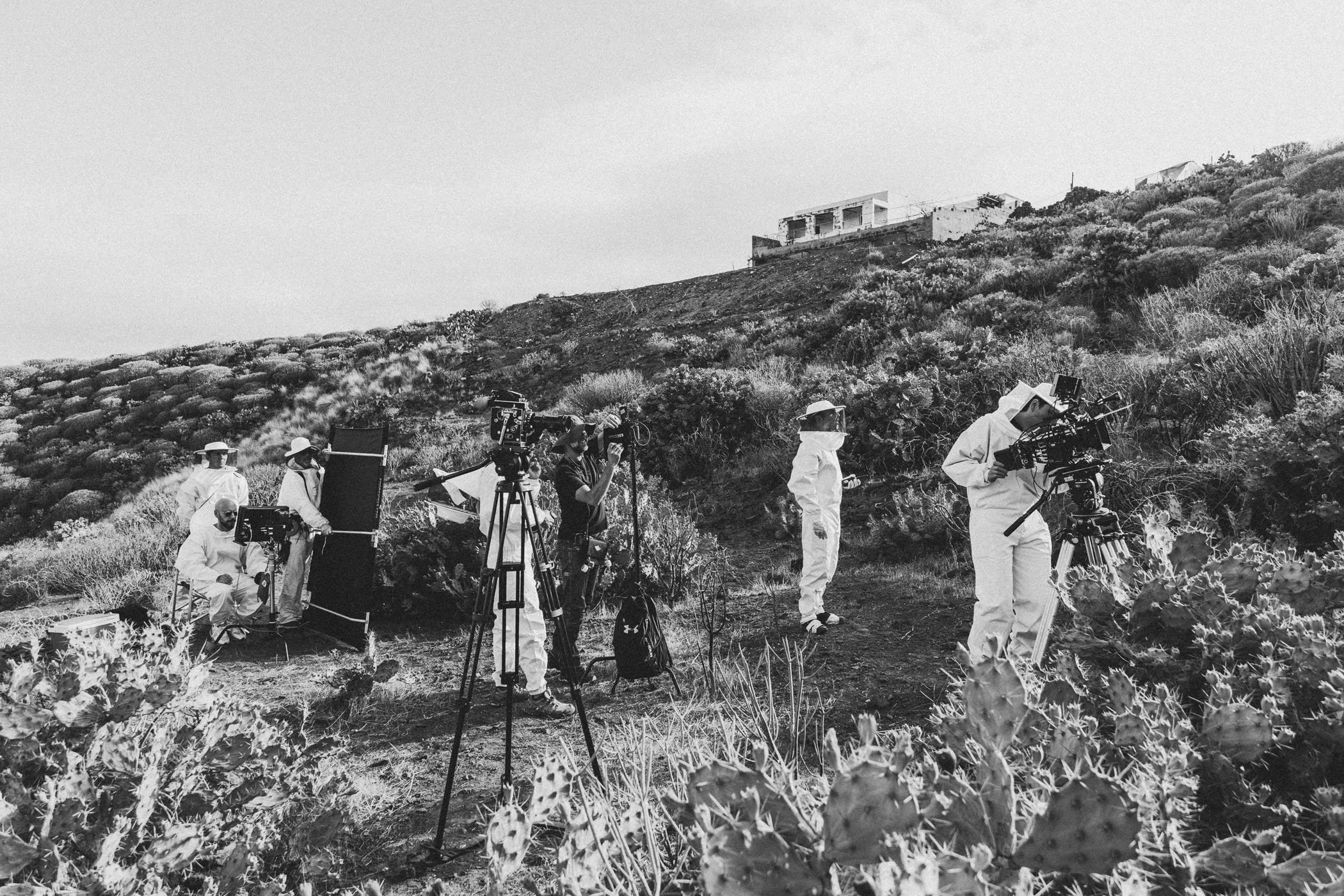 Equipo de filmación en un entorno árido con cactus y vegetación, con cámaras y personas con equipo de protección frente a un paisaje montañoso y una estructura en la cima.