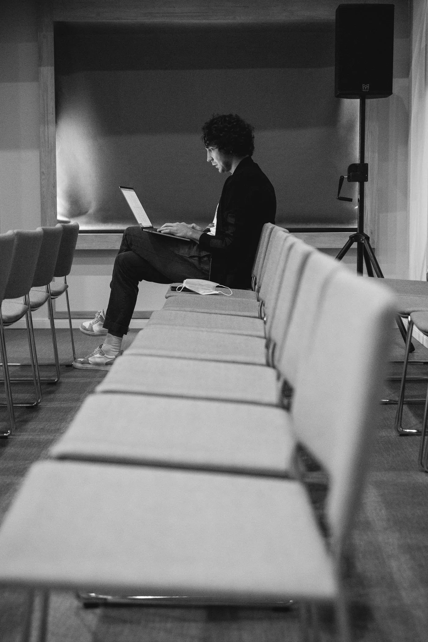 Persona sentada en una sala de conferencias con sillas vacías, usando una laptop, con una mascarilla sobre la mesa y en un entorno de interior