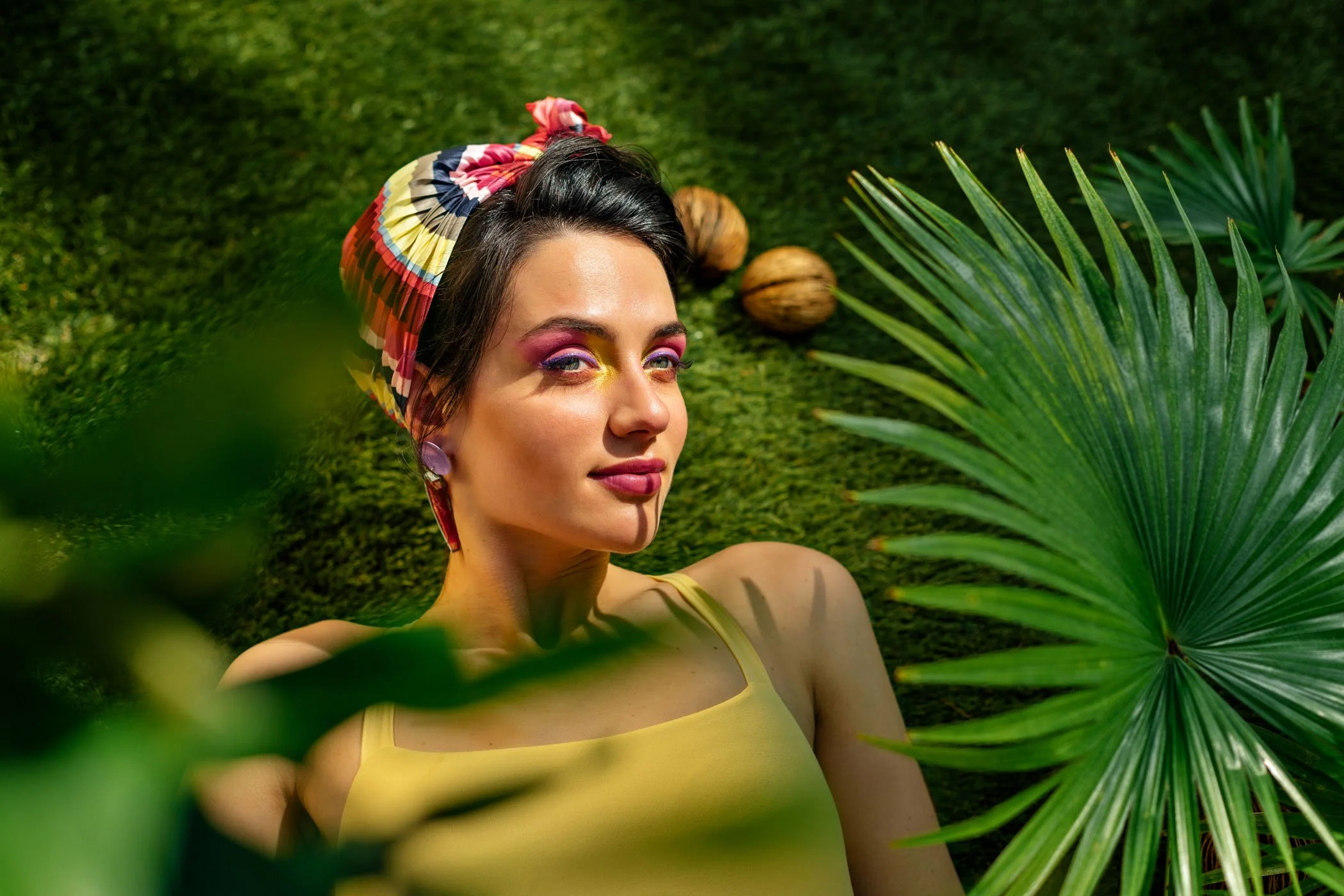 Mujer con maquillaje colorido, usando una pañoleta con patrón vibrante, rodeada de plantas tropicales y cocos en un entorno natural.