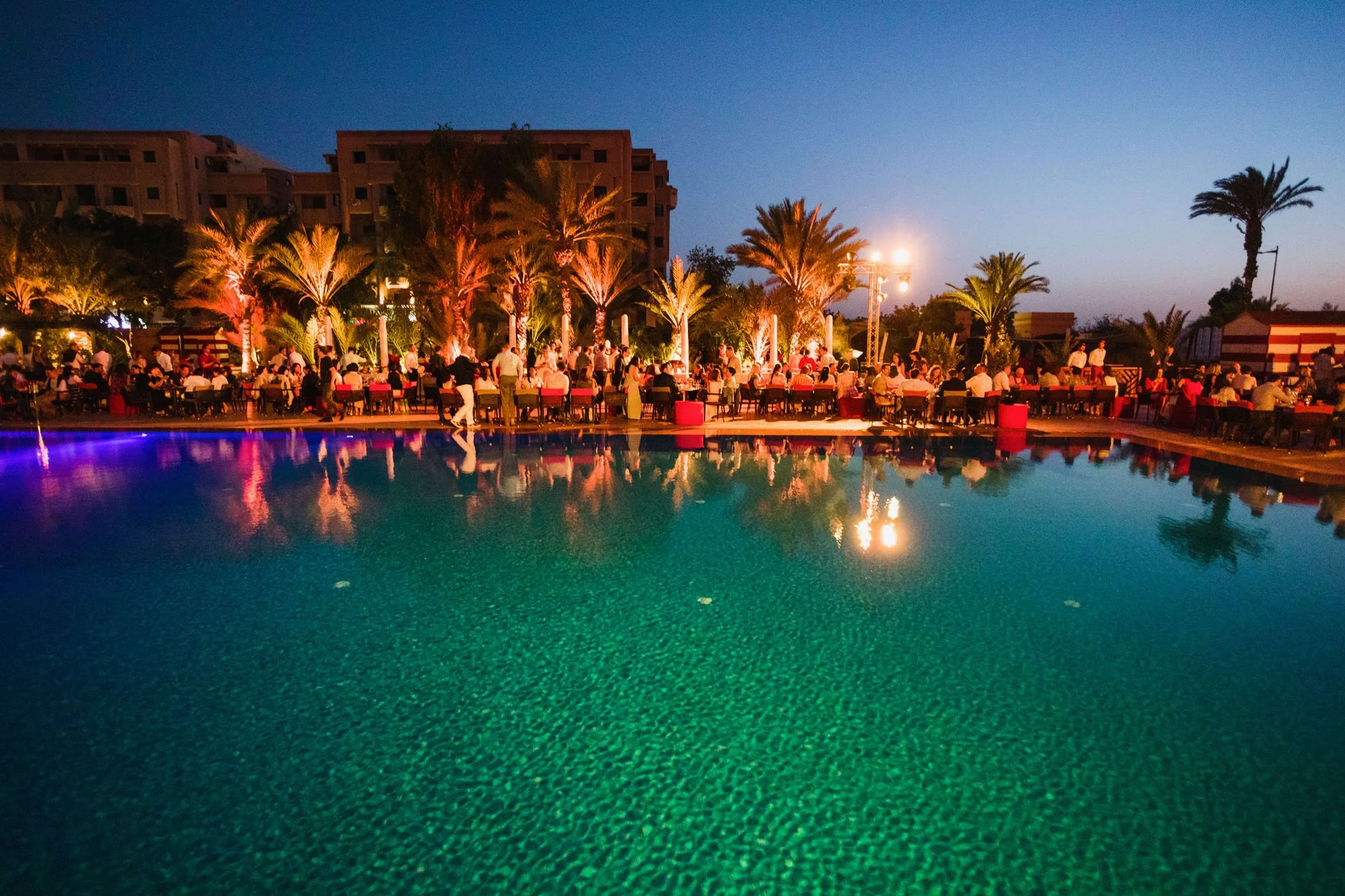 Una piscina con agua azul y una multitud de personas en una terraza al aire libre con palmeras, iluminación cálida y mesas, en la noche.