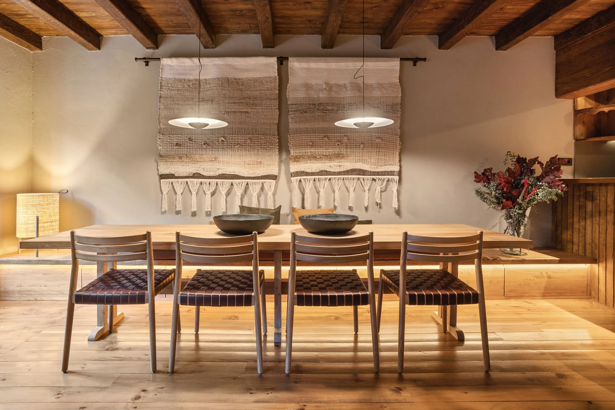 Un comedor con mesa de madera y seis sillas, decoración con arte textil en la pared, lámparas colgantes y una lámpara de escritorio, con una planta con flores en el lado derecho.