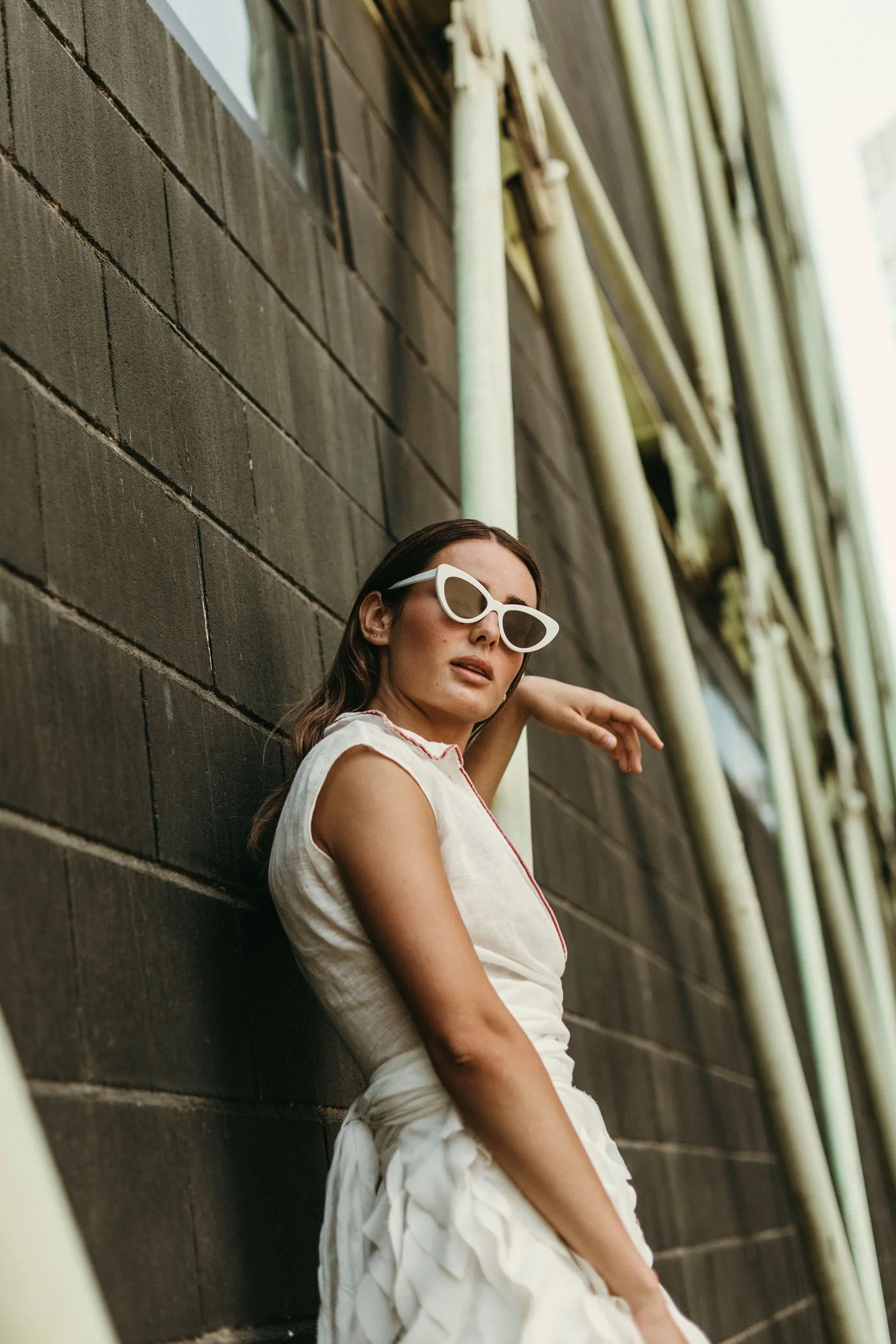 Mujer usando gafas blancas y vestido blanco, apoyada contra una pared de ladrillos oscuros, con caños amarillos en el fondo.