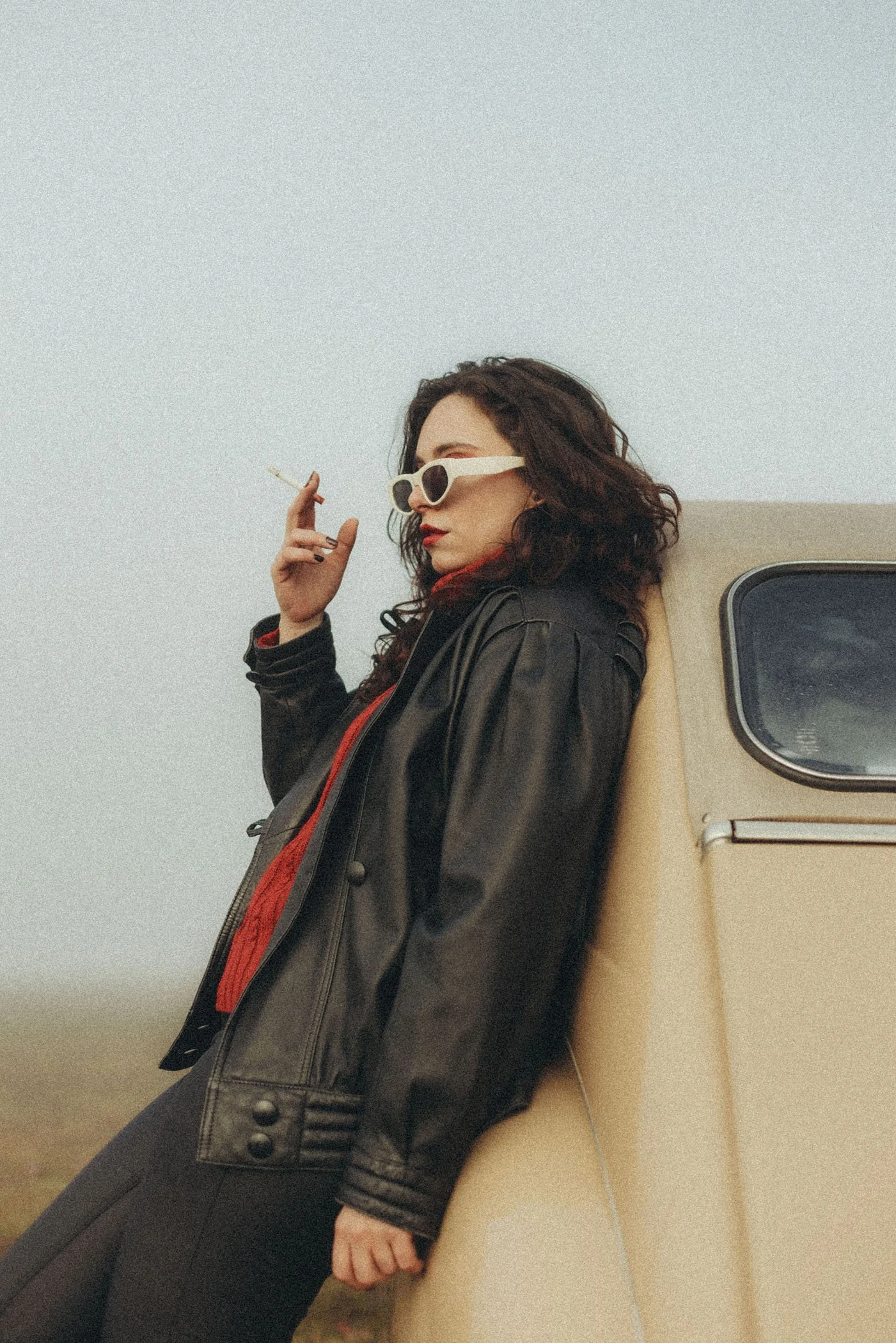 Mujer con gafas blancas, cabello rizado, vestida con chaqueta de cuero negra, apoyada en una máquina amarilla, sosteniendo un cigarrillo, en un ambiente exterior y despejado.