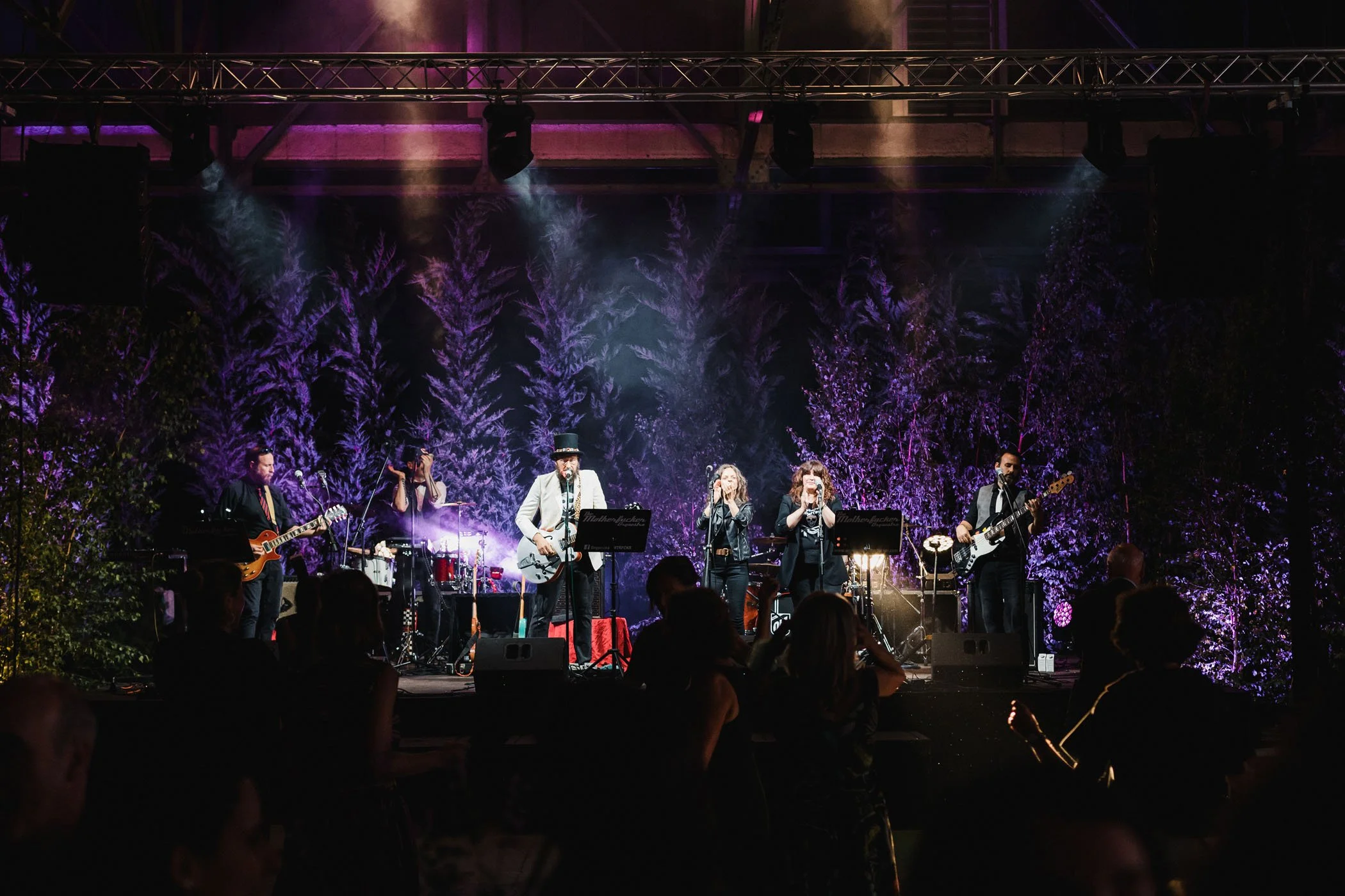 Conjunto musical tocando en escenario iluminado con luces moradas y fondo de árboles, público viendo la presentación.