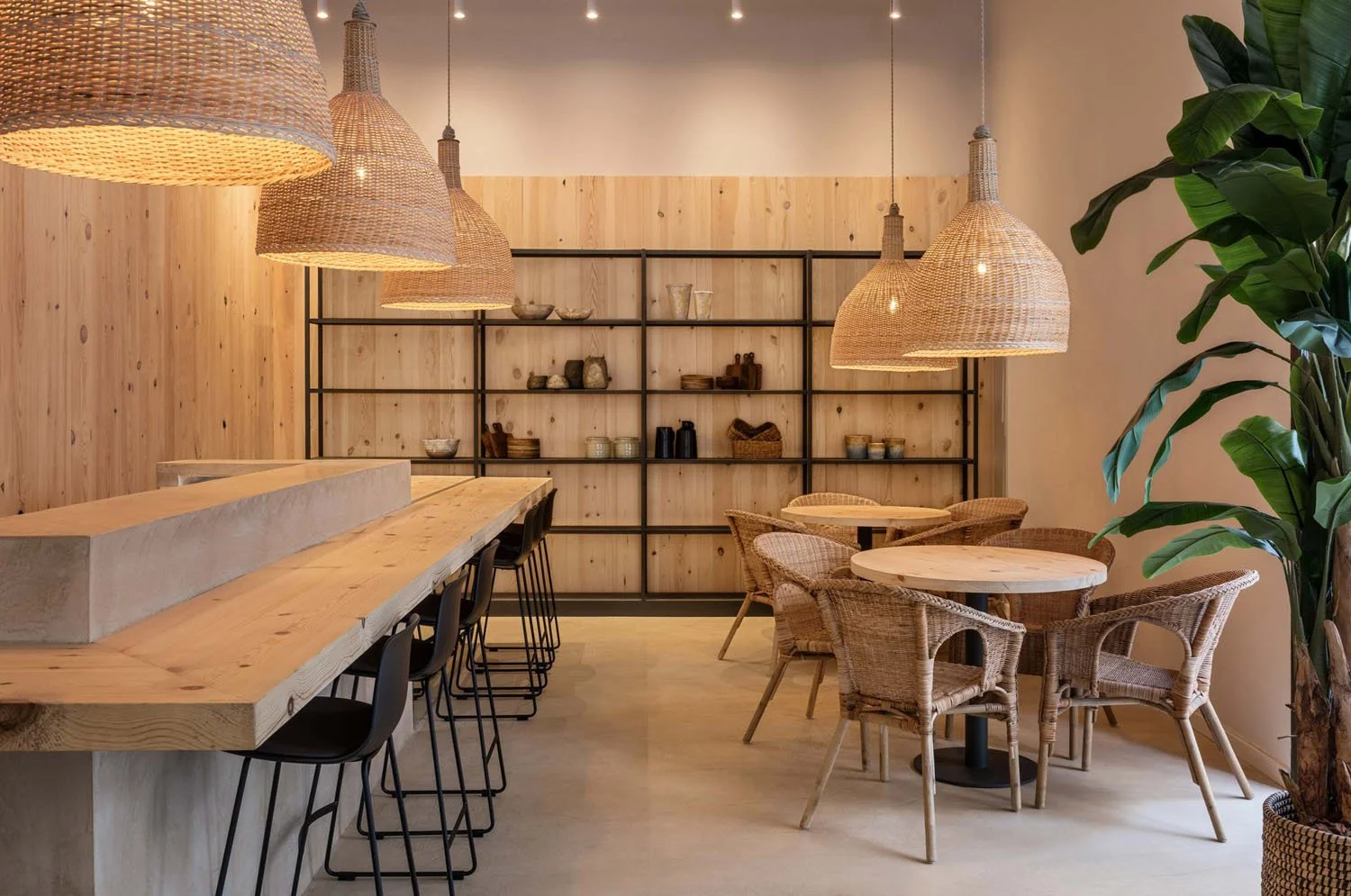 Interior de un café con decoracion de madera, sillas de mimbre, mesas redondas de madera, estantería negra con objetos decorativos y un árbol grande.