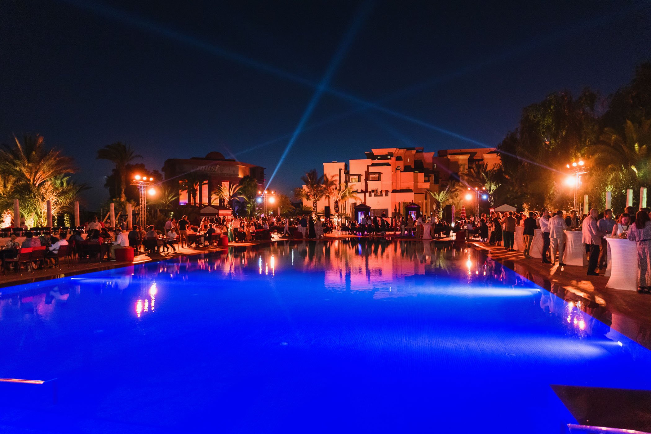 Fiesta en una piscina iluminada con luces de colores, con personas disfrutando, escenario con luces y estructuras modernas en el fondo, rodeado de palmeras y árboles