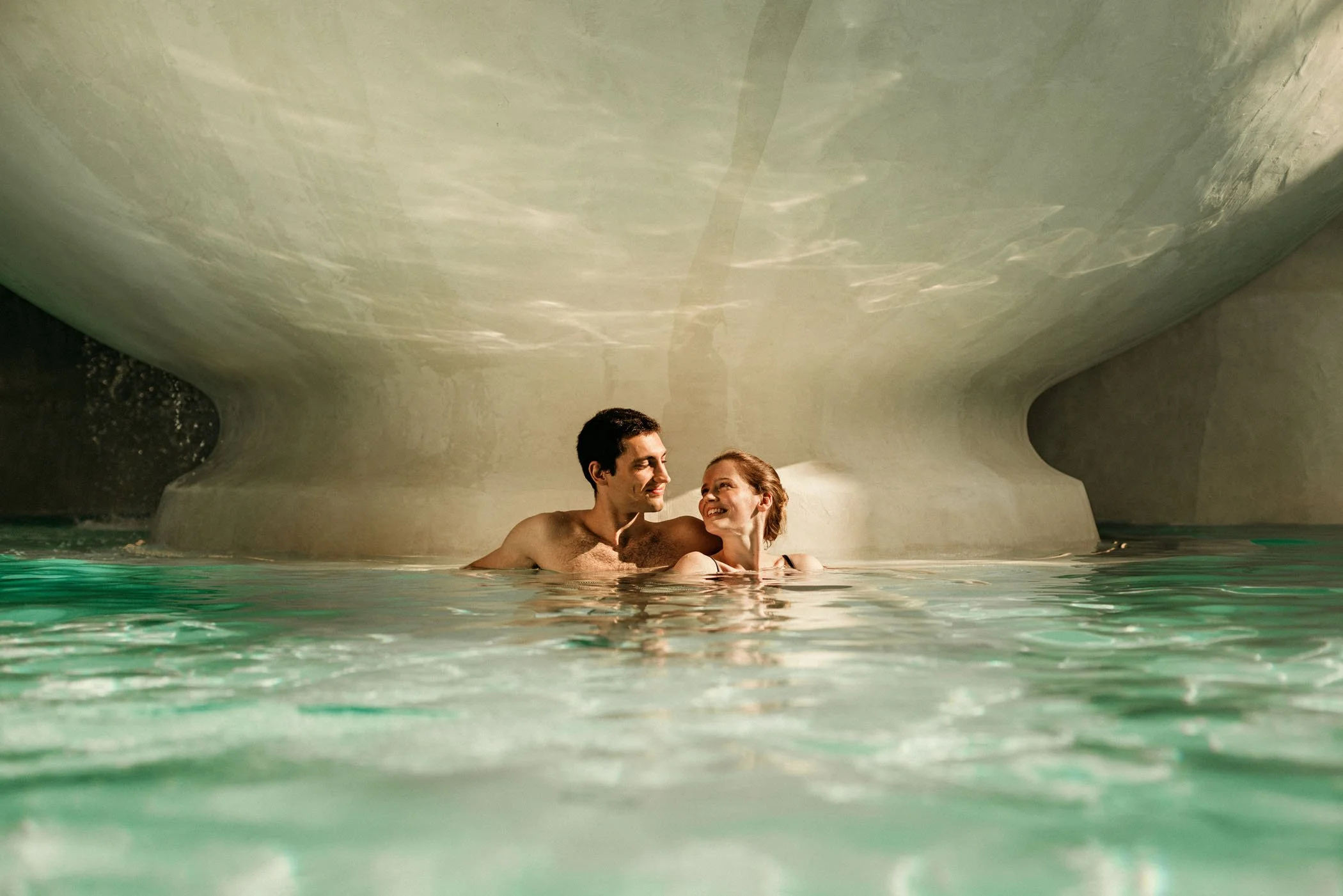 Una pareja disfrutando en una piscina cubierta, con la estructura del techo visible en el fondo.