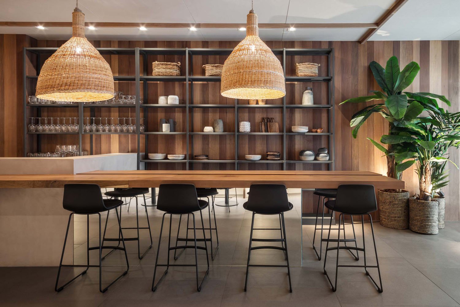 Interior de restaurante con estanterías de madera y metal con platos y vasos, barra de madera y sillones negros, lámparas de mimbre y plantas grandes en cestas de mimbre.