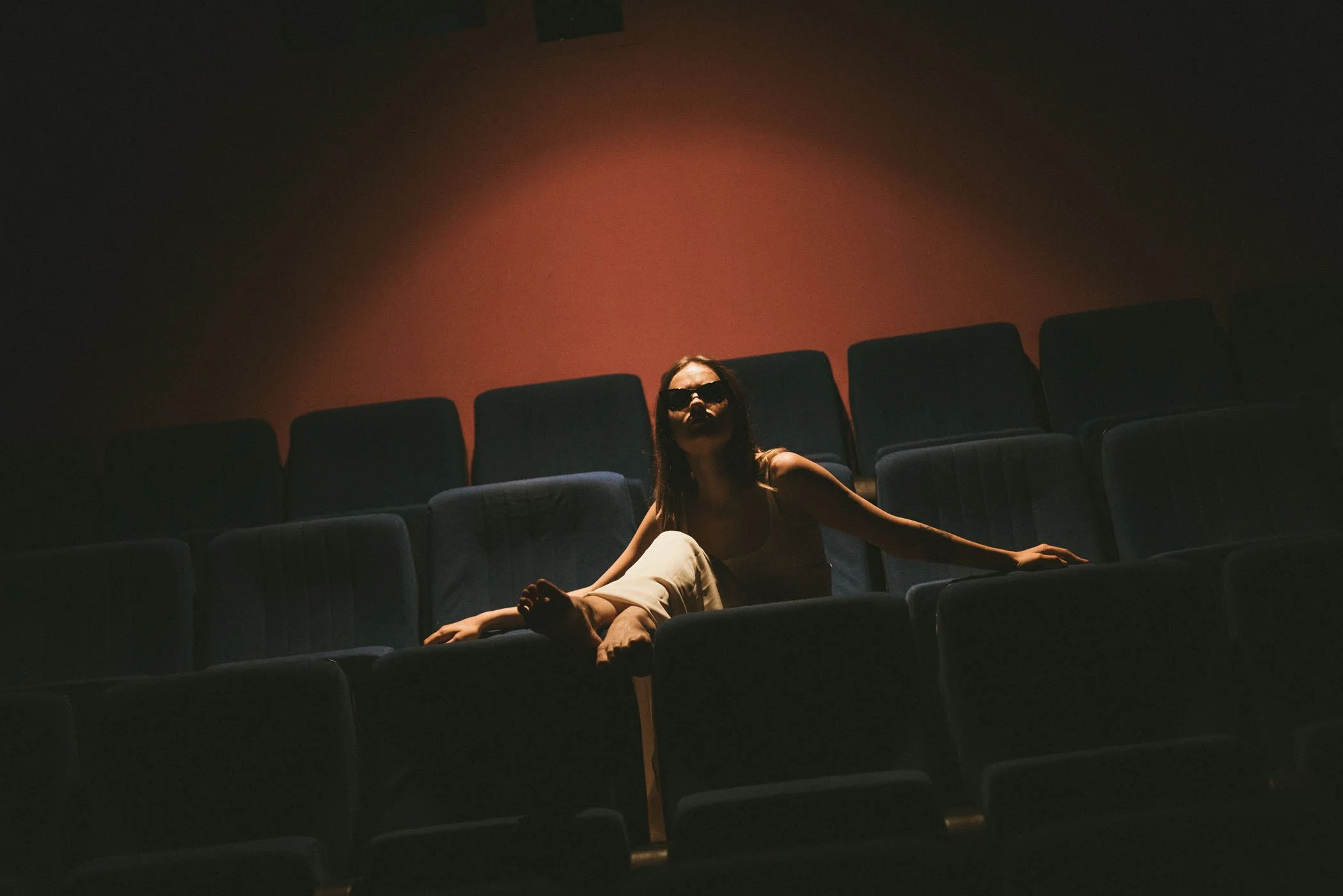 Mujer con gafas oscuras sentada en una sala de cine oscura, con butacas azules, iluminada por una luz suave y tenue.