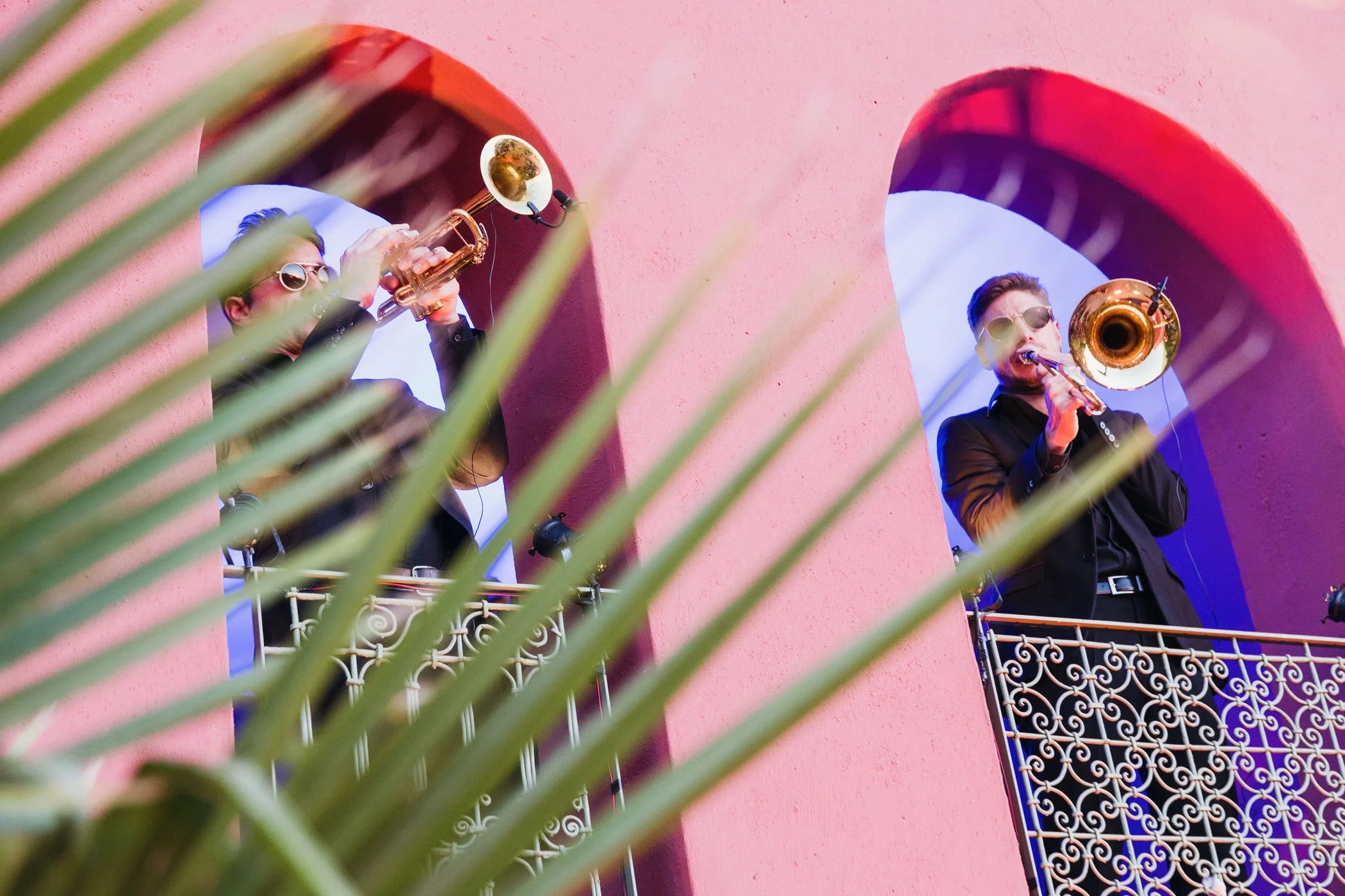 Dos músicos tocando trompeta en un balcón frente a un fondo de paredes rosadas y arcos blancos, con hojas verdes en primer plano.
