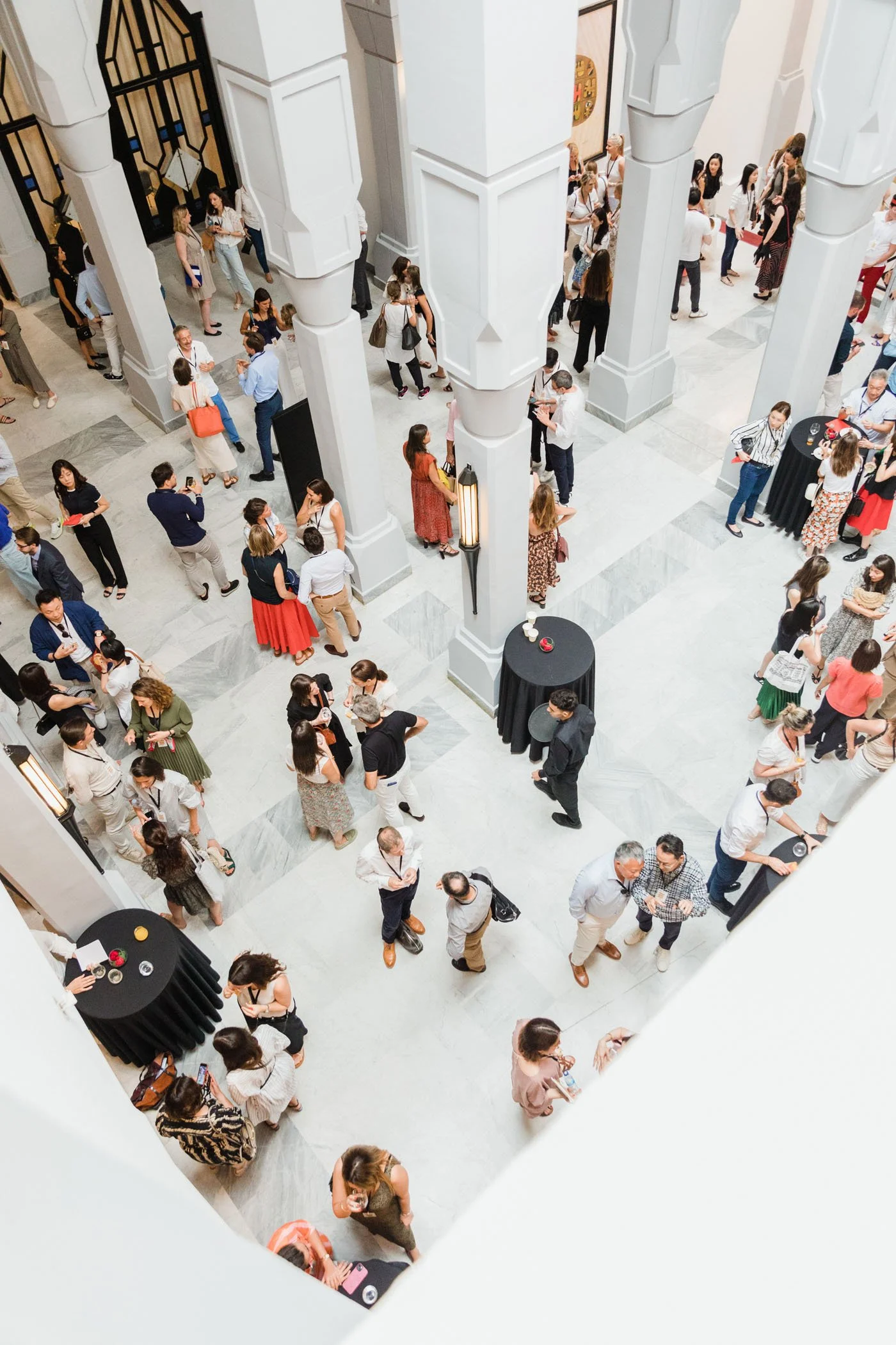 Vista aérea de una multitud de personas en un evento en un edificio con arquitectura moderna, con columnas y paredes blancas, y mesas con manteles negros.