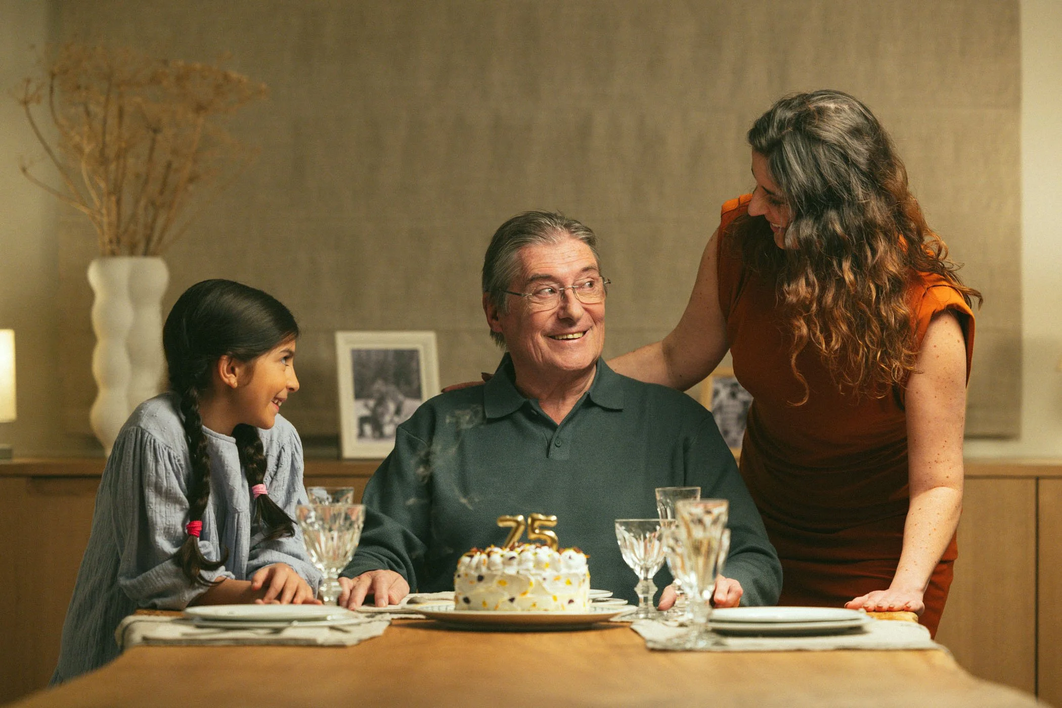 Una familia celebrando el 75 aniversario, con un pastel decorado en la mesa, dos adultos y una niña sonriendo y compartiendo un momento feliz en la cena.