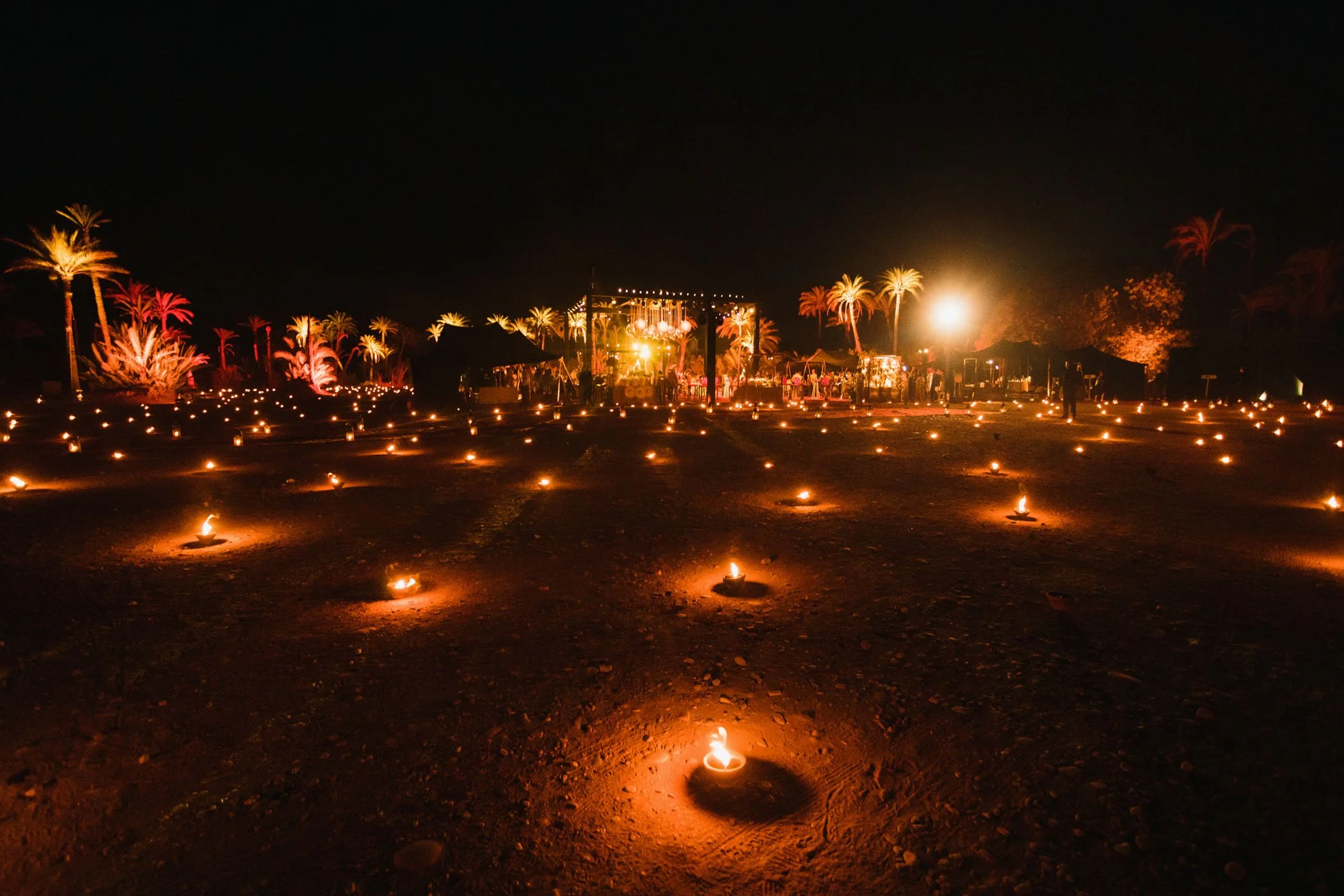 Escenario de noche iluminado con velas en la arena, palmeras decoradas con luces y una tarima de eventos en el fondo.