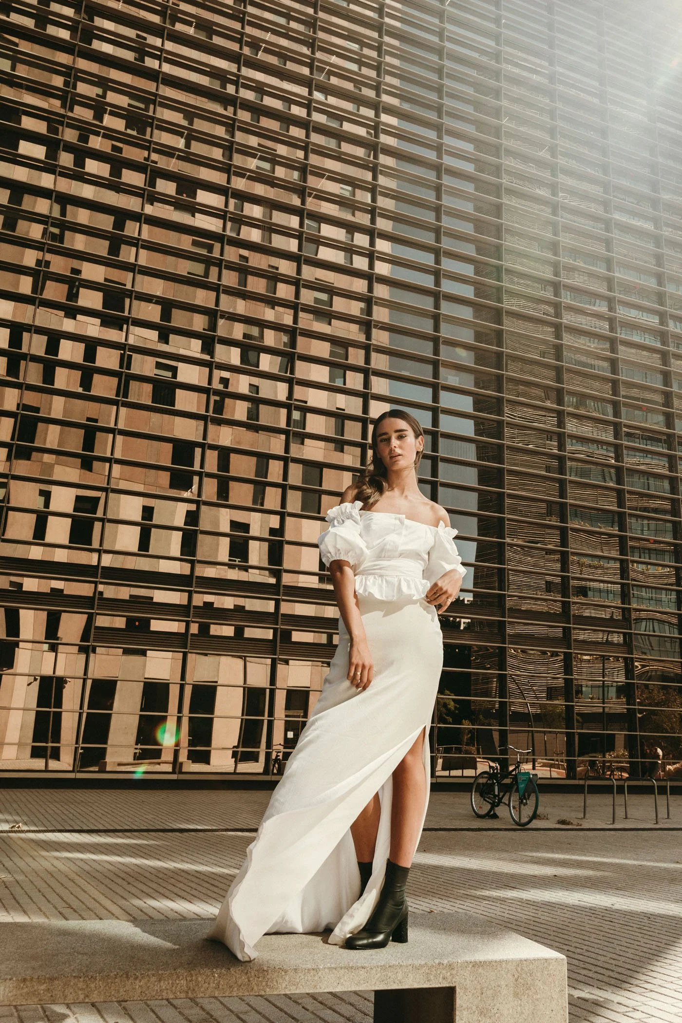 Mujer posando en ciudad con edificio de fondo, usando vestido blanco con abertura y botas negras