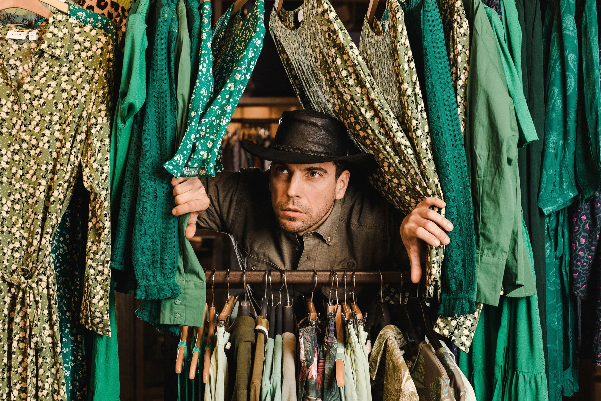 Hombre con sombrero negro entre ropa de colores verdes y estampados, mirando hacia adelante mientras mira por una cortina de ropa en una tienda de ropa.