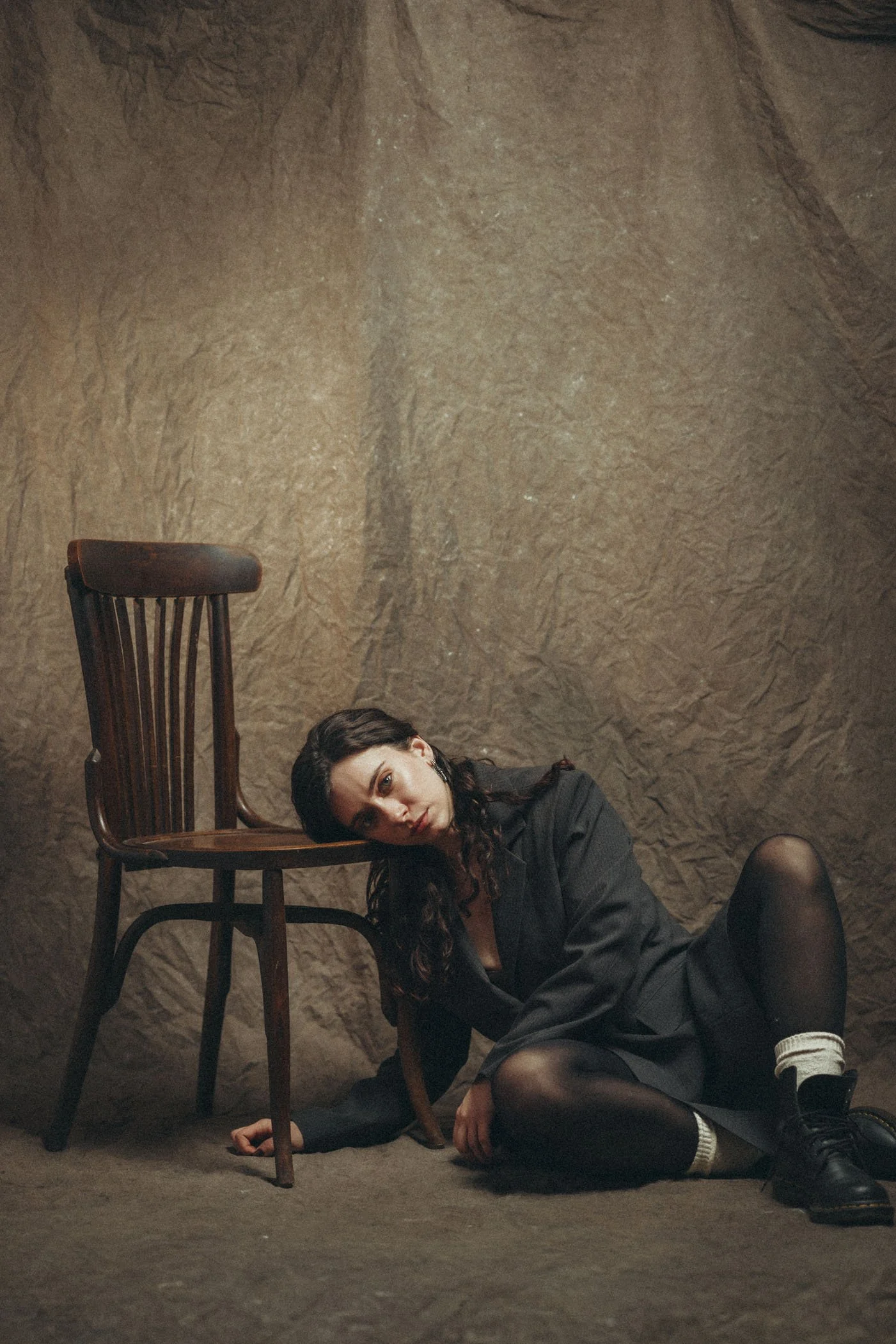 Una mujer con ropa oscura, sentada en el suelo cerca de una pared de piedra, apoyada en una silla de madera