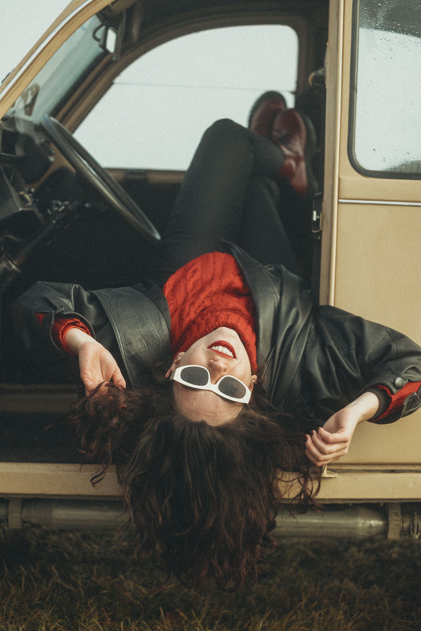 Mujer con gafas blancas acostada sobre el suelo y apoyada en la puerta abierta de un vehículo antiguo, vistiendo chaqueta de cuero y bufanda roja.