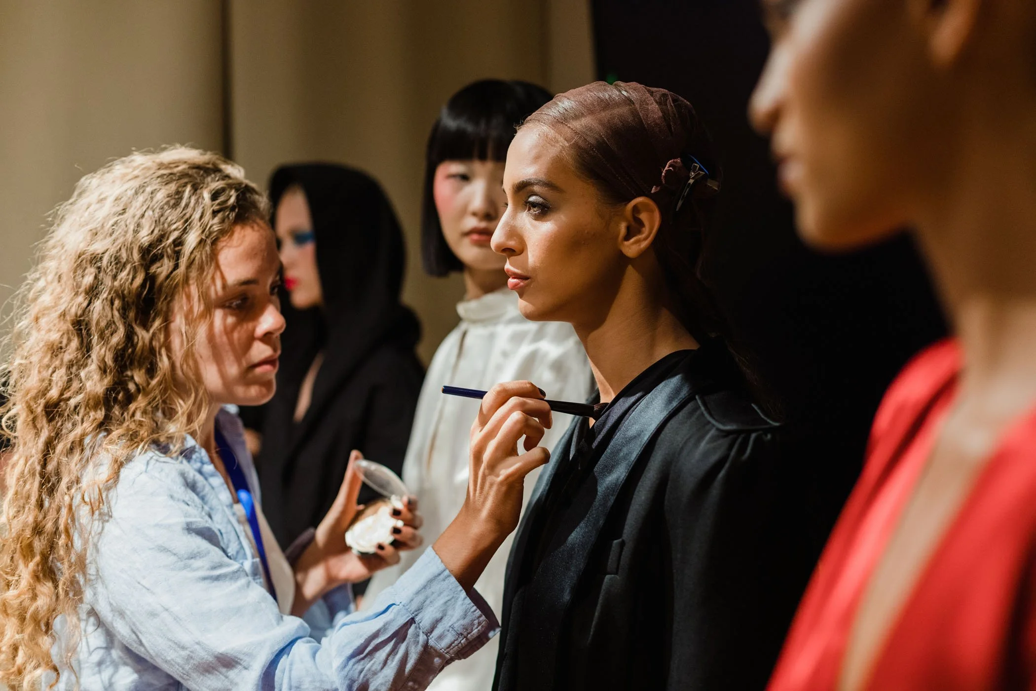 Una mujer con maquillaje y cabello en un peinado elegante, en un evento, recibe ayuda de otra mujer con cabello rizado y rubio, mientras otras chicas con diferentes estilos de cabello observan.