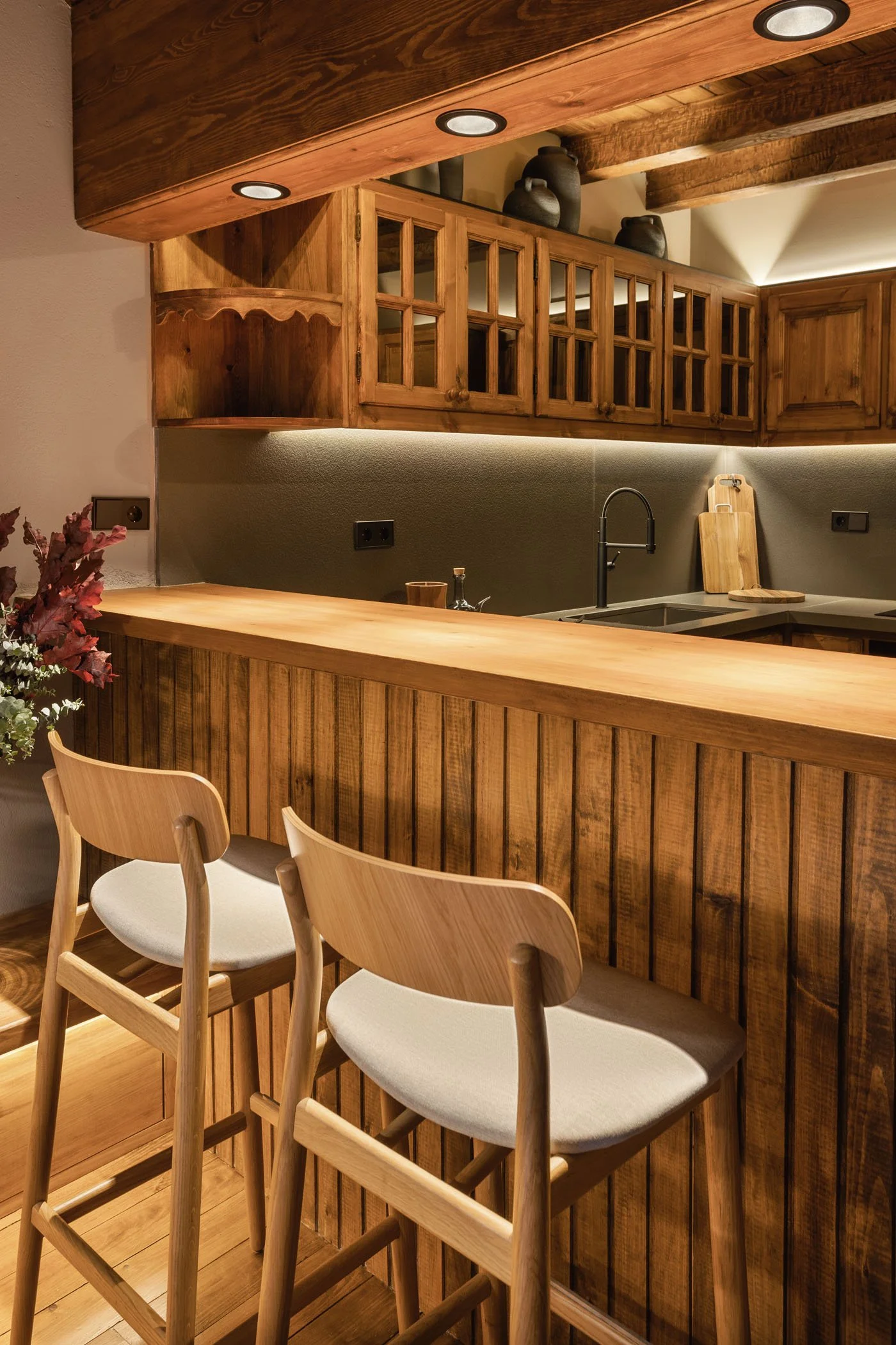 Barra de cocina de madera con dos sillas de madera claras y tapizadas en blanco, armarios superiores y una encimera de madera, con utensilios y adornos negros, en un ambiente cálido y acogedor.