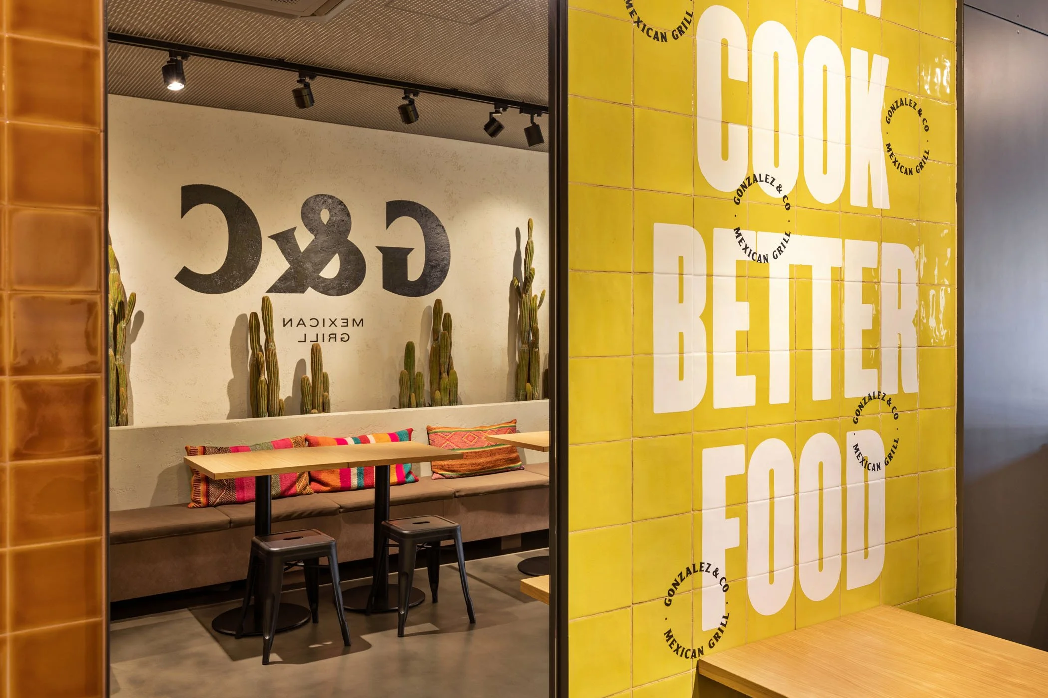 Vista de un restaurante mexicano llamado Gonzlez & Co con mesas, sillas y decoraciones de cactus, paredes decoradas con letras grandes que dicen 'COM BEBER' y 'GONZALEZ & CO MEXICAN GRILL'.