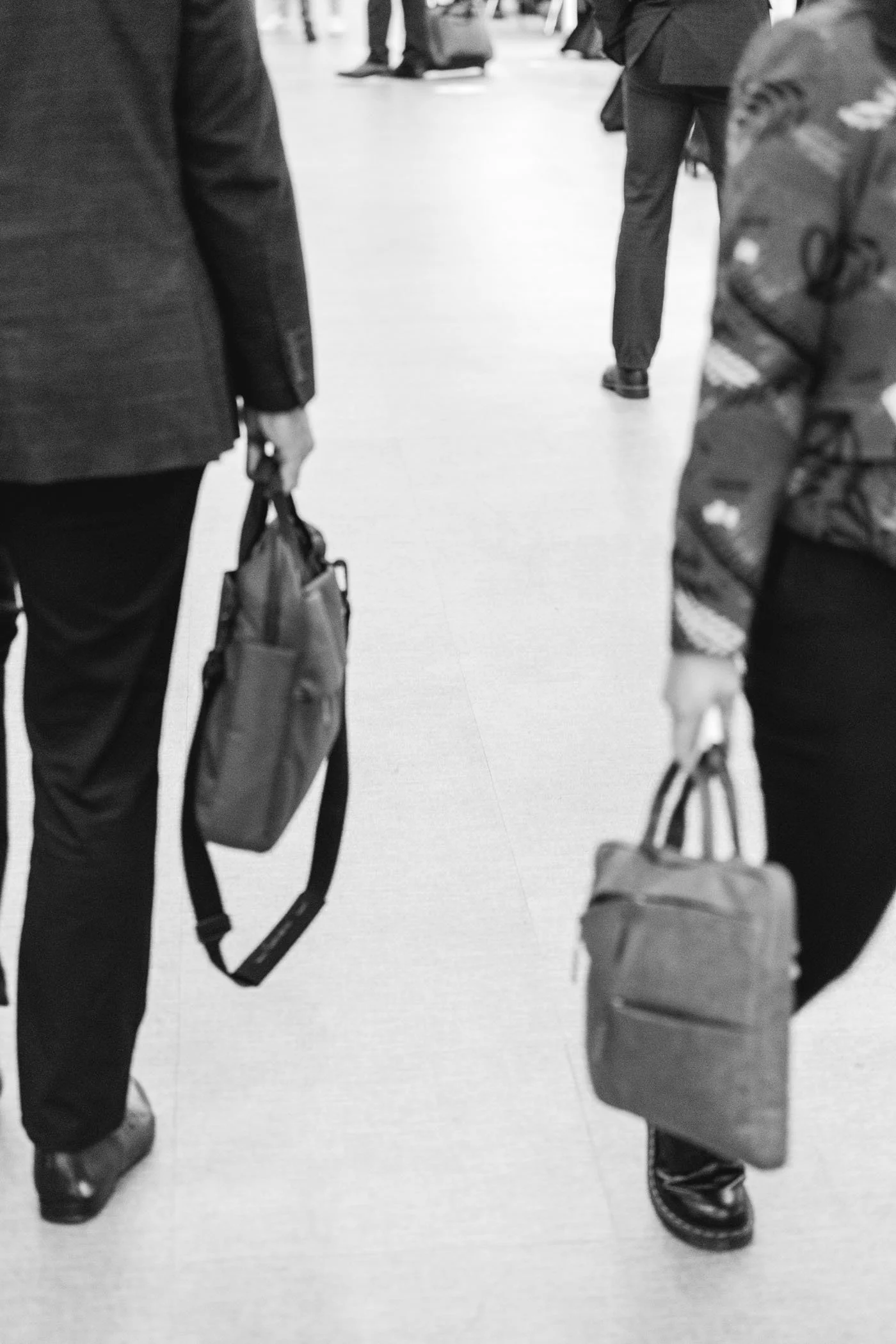 Vista en un aeropuerto con dos personas de espalda, ambas llevan maletines, una con bolso y la otra con mochila. La escena es en blanco y negro.