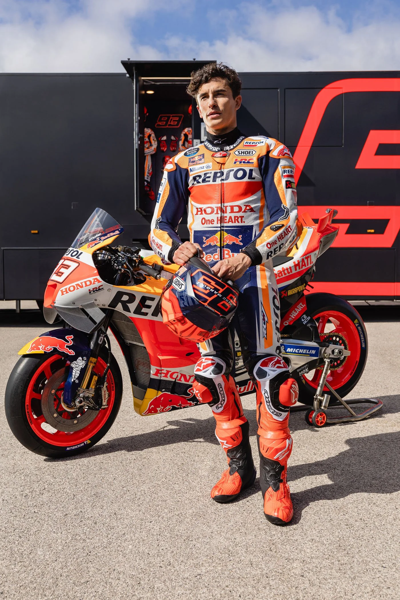 Piloto de motocicleta en traje y botas de carreras, sosteniendo casco, en pista de carreras con motocicleta de carrera patrocinada por Repsol y Honda, y remolque de transporte en el fondo.