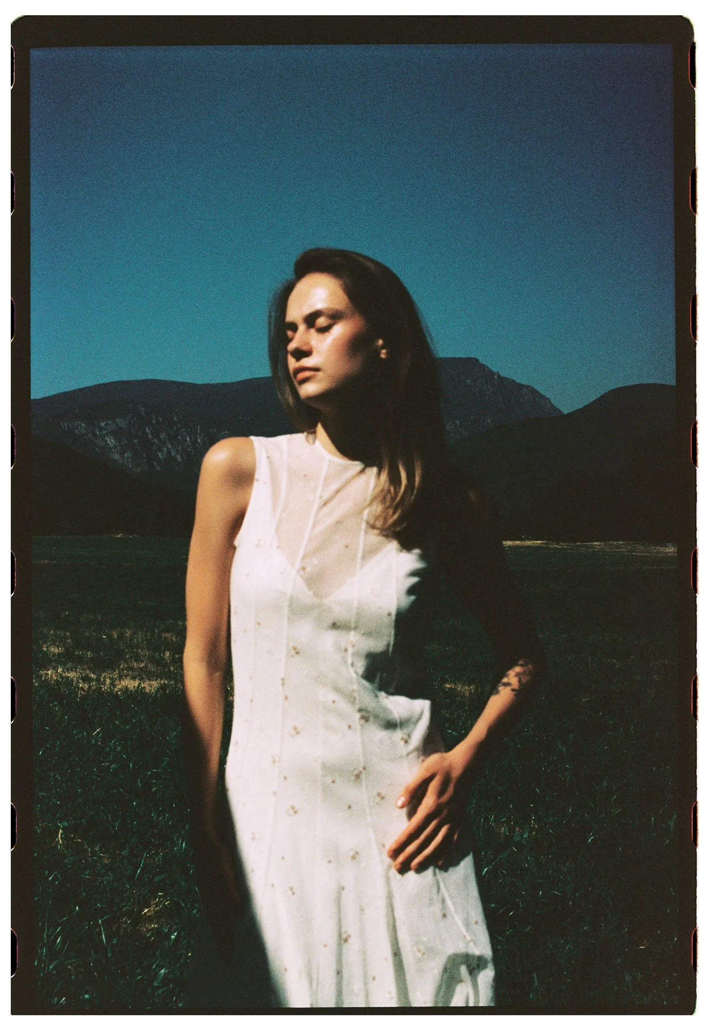 Una mujer con vestido blanco de pie en un campo con montañas y un cielo azul oscuro en el fondo.