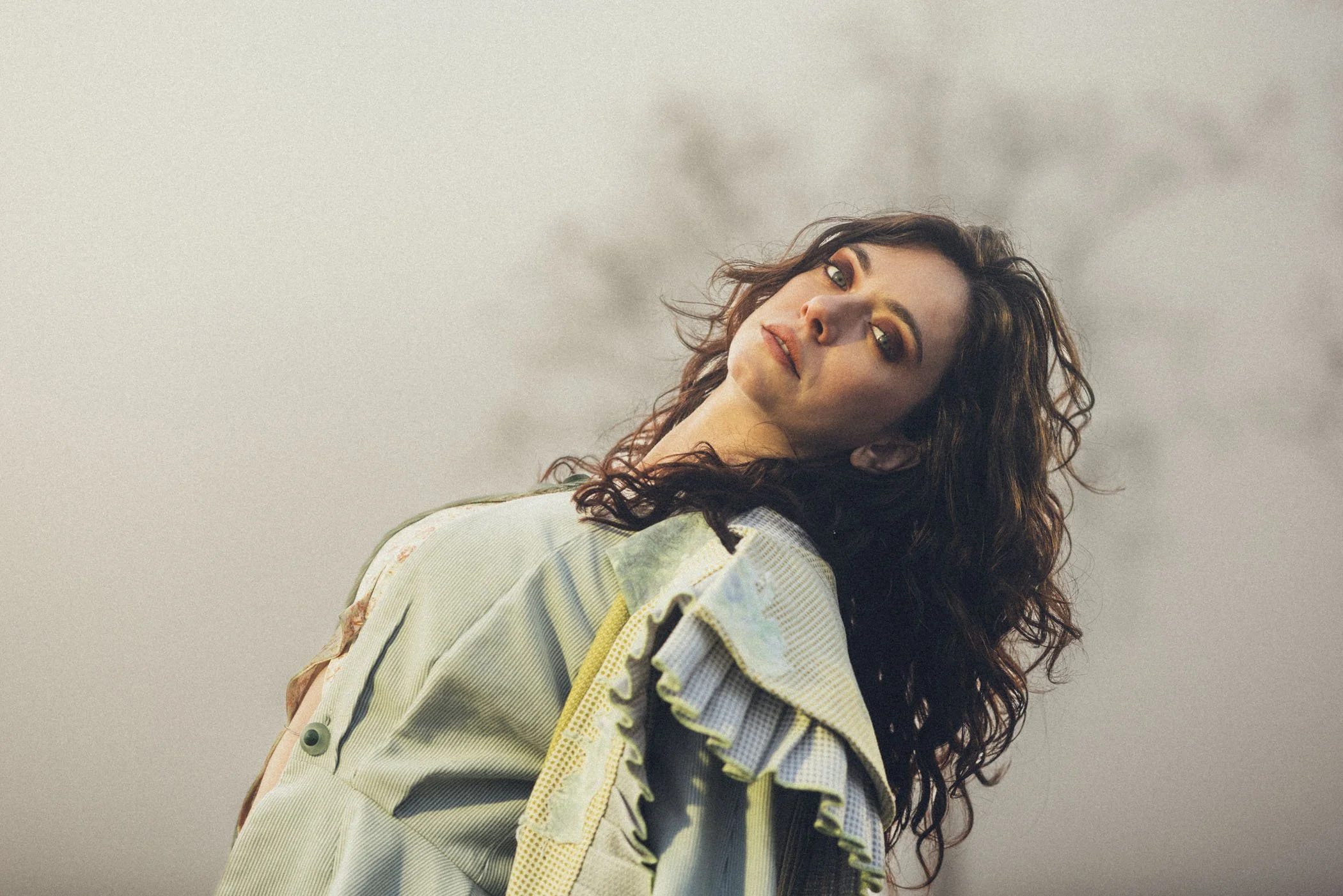 Retrato de una mujer con cabello rizado y maquillaje en tonos dorados, mirando hacia la cámara, con un fondo grisáceo.