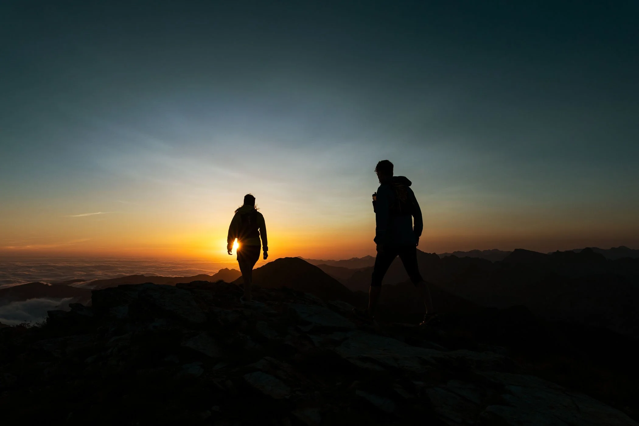 Dos personas caminando en la cima de una montaña durante un atardecer o amanecer, con un cielo con colores cálidos y un paisaje montañoso en el fondo.