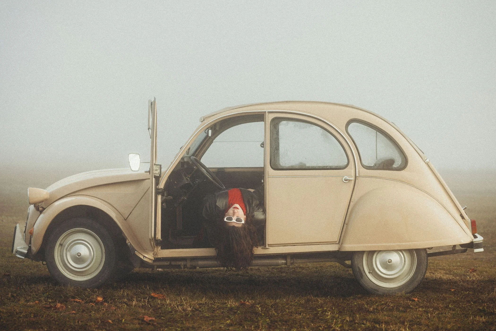 Una mujer con gafas blancas colgando del lado abierto de un coche vintage beige en un campo con niebla.
