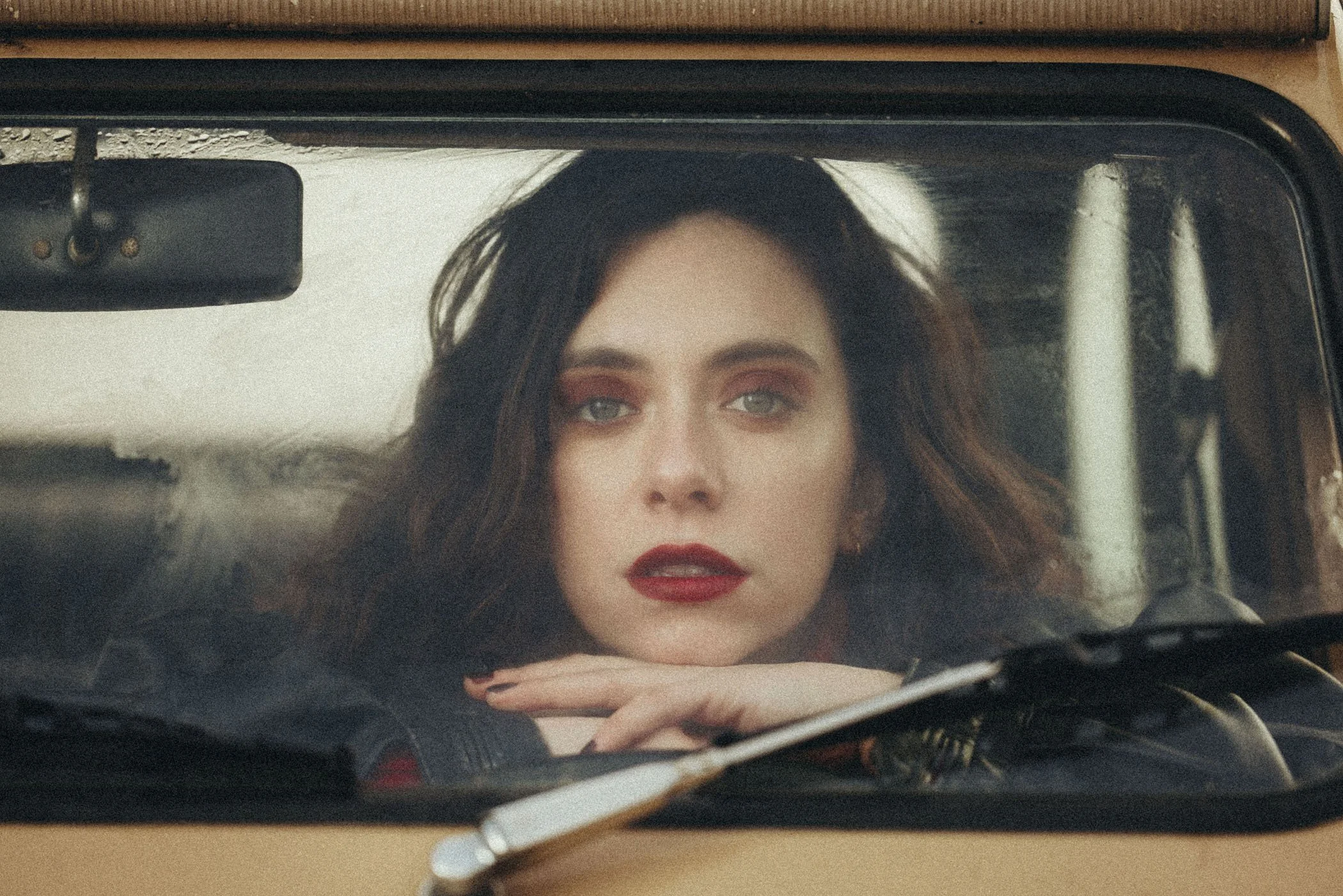 Mujer con cabello castaño oscuro y maquillaje de labios rojos, observando a través del parabrisas de un automóvil clásico.