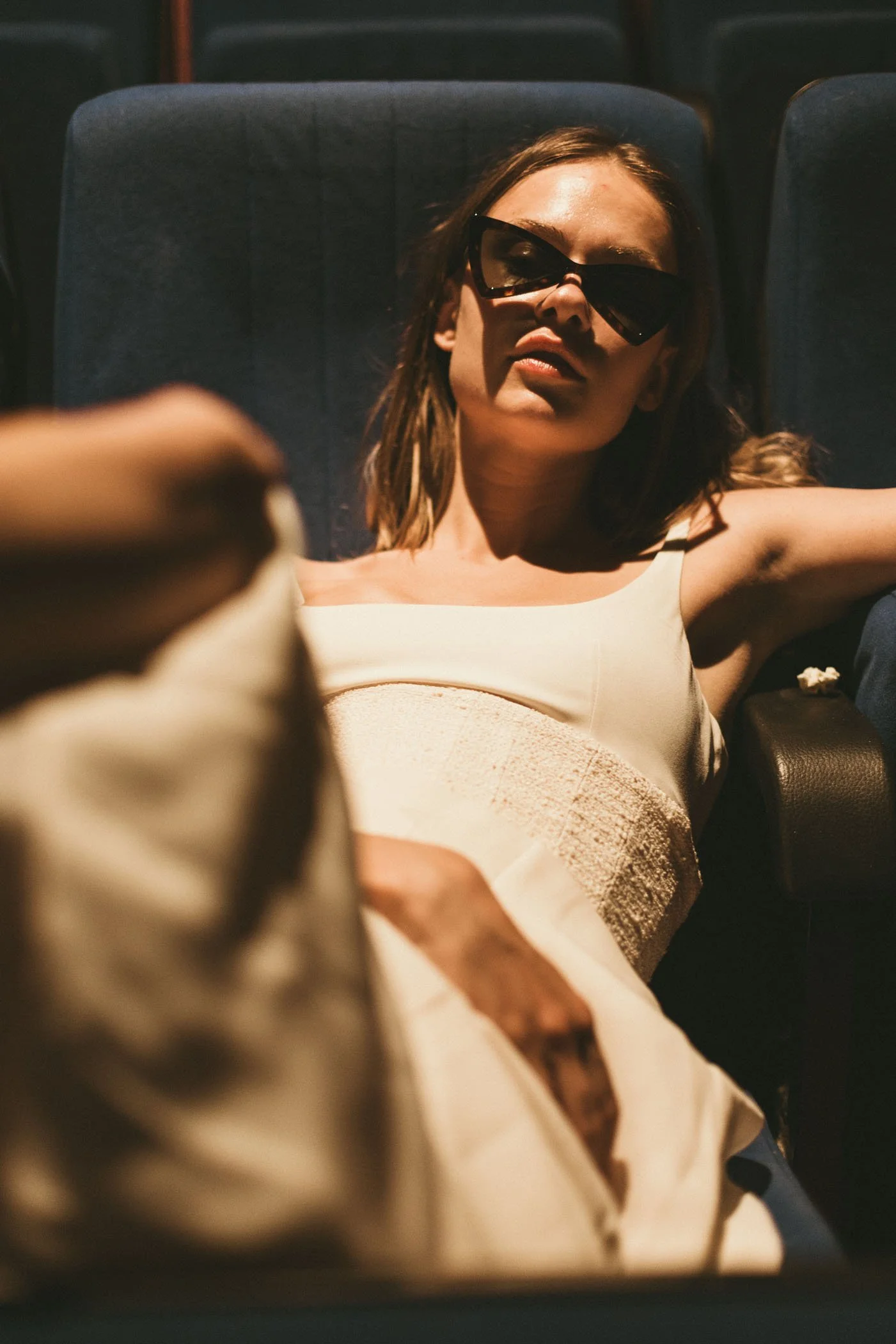 Mujer joven con gafas de sol acostada en un asiento de cine, vestida con ropa de color claro.