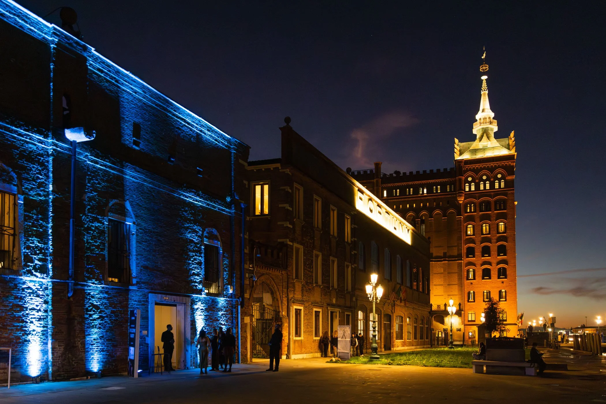Vista nocturna de un edificio histórico iluminado con luces azules y amarillas, incluyendo una torre con punta dorada, en un entorno urbano con personas caminando y bancos en primer plano.