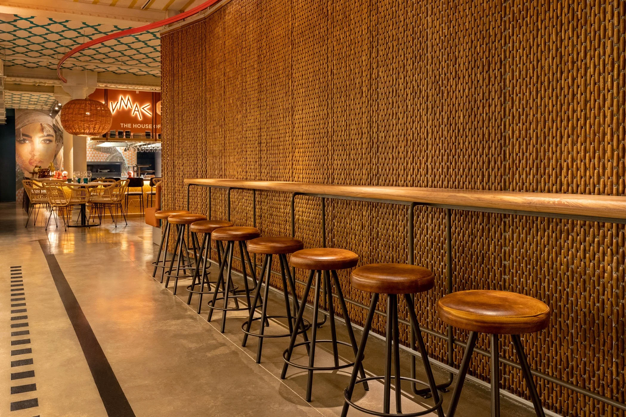 Interiores de un restaurante con una fila de seis taburetes de madera junto a una pared de ratán, en el fondo mesas decoradas y un cartel que dice 'MAMÁGICA'.