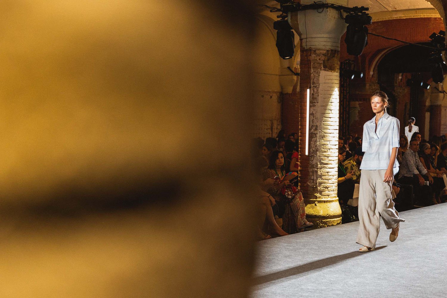 Modelo caminando en pasarela durante una fashion show en un lugar con arquitectura de estilo antiguo, con audiencia sentada a los lados y luces enfocadas en la pasarela.