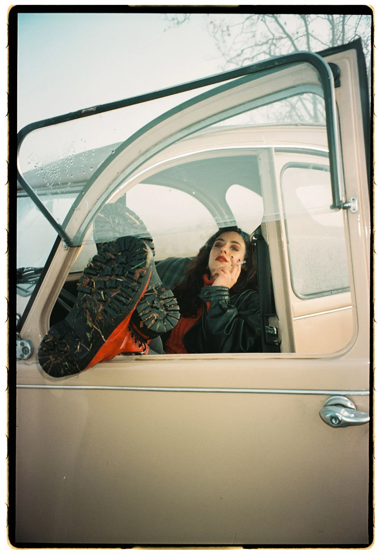 Mujer sentada en la cabina de un vehículo clásico, con la pierna levantada y tocándose la cara, en un día nublado y con árboles desnudos en el fondo.