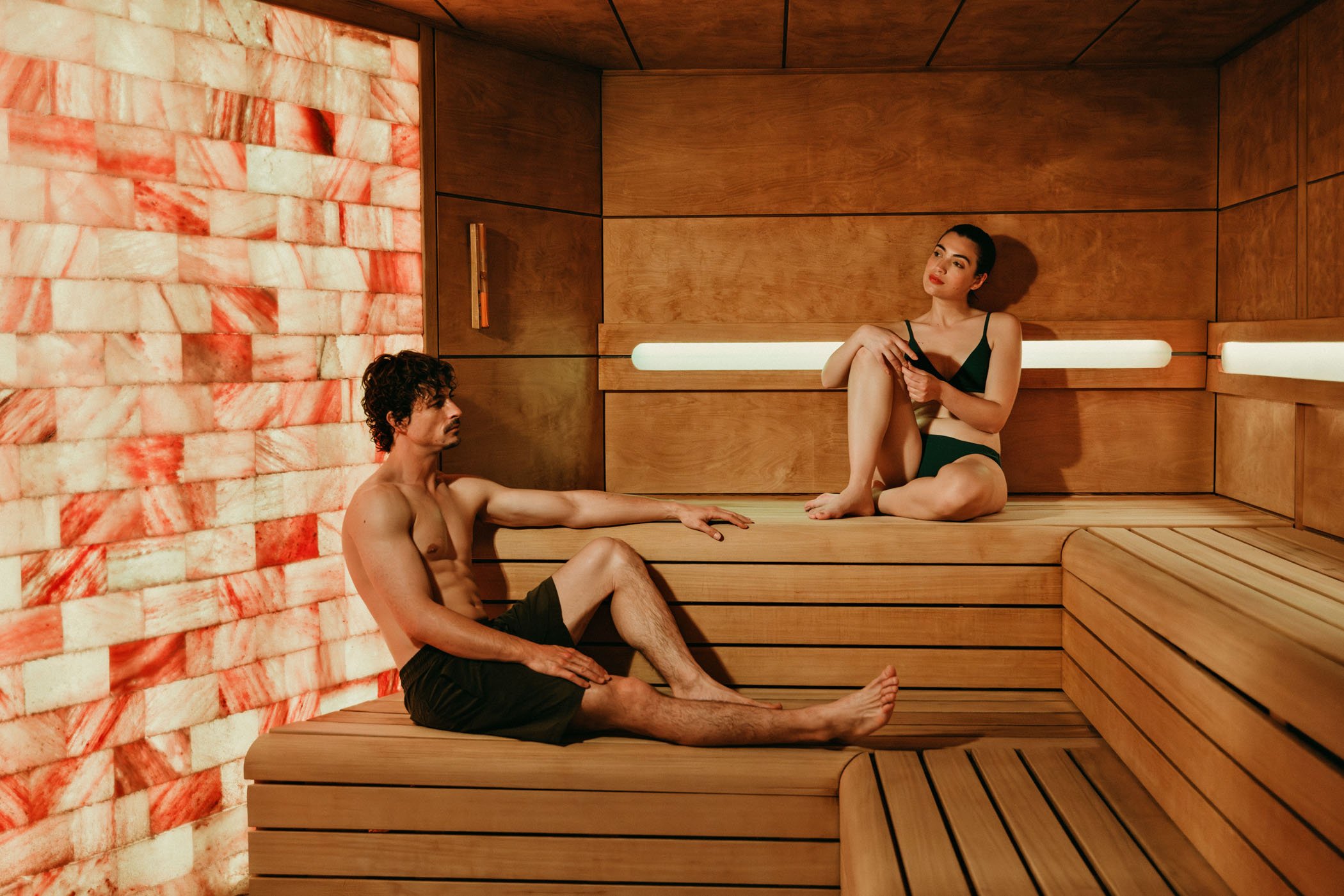 Una pareja en una sauna de madera, el hombre está sentado con las piernas extendidas y la mujer está sentada con las piernas dobladas, ella se mira pensativa mientras sostiene una taza.