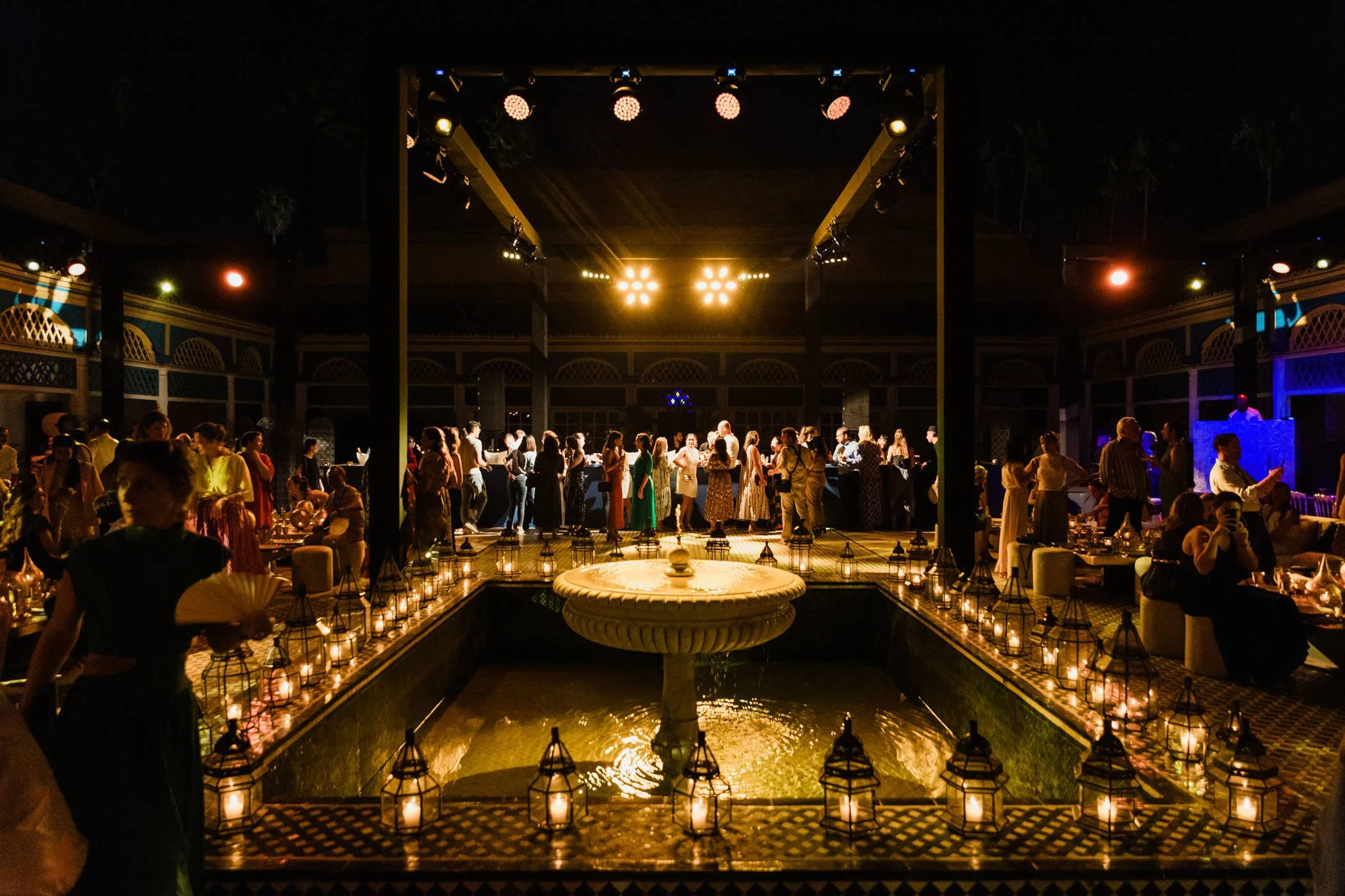Imagen de una fiesta en un patio decorado con velas, con muchas personas bailando y conversando, en el fondo hay una área de barra y iluminación cálida.
