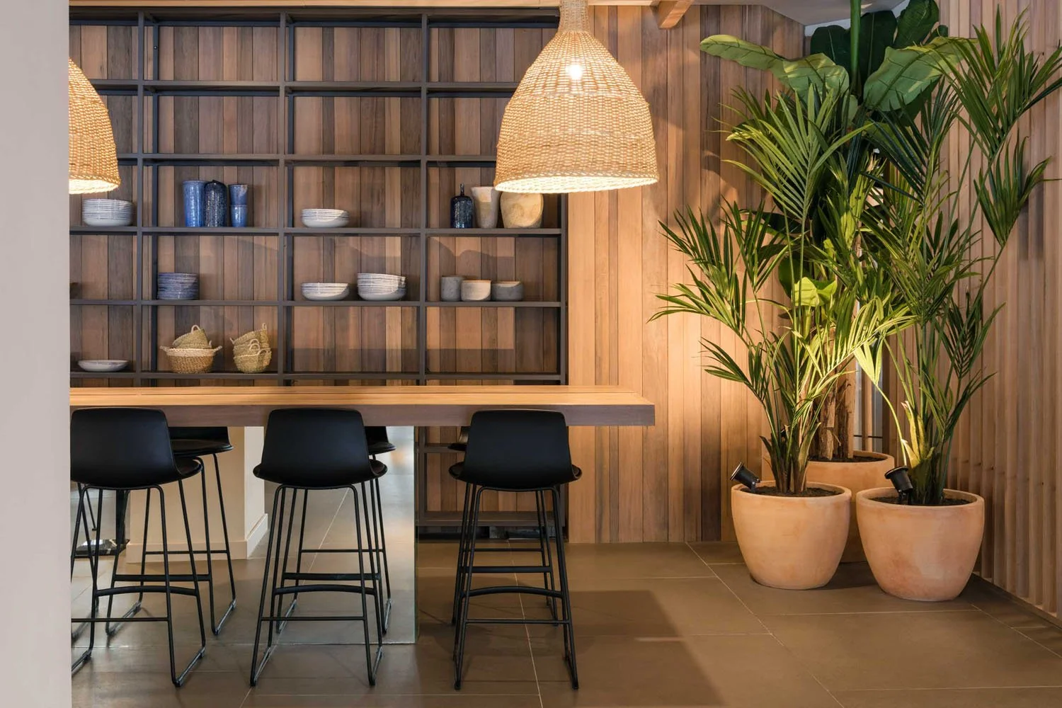 Interiores de una área de comedor con estantes de madera con platos y tazas, 3 sillas negras, una mesa de bar de madera, lámparas de mimbre colgantes y plantas verdes en macetas de barro.