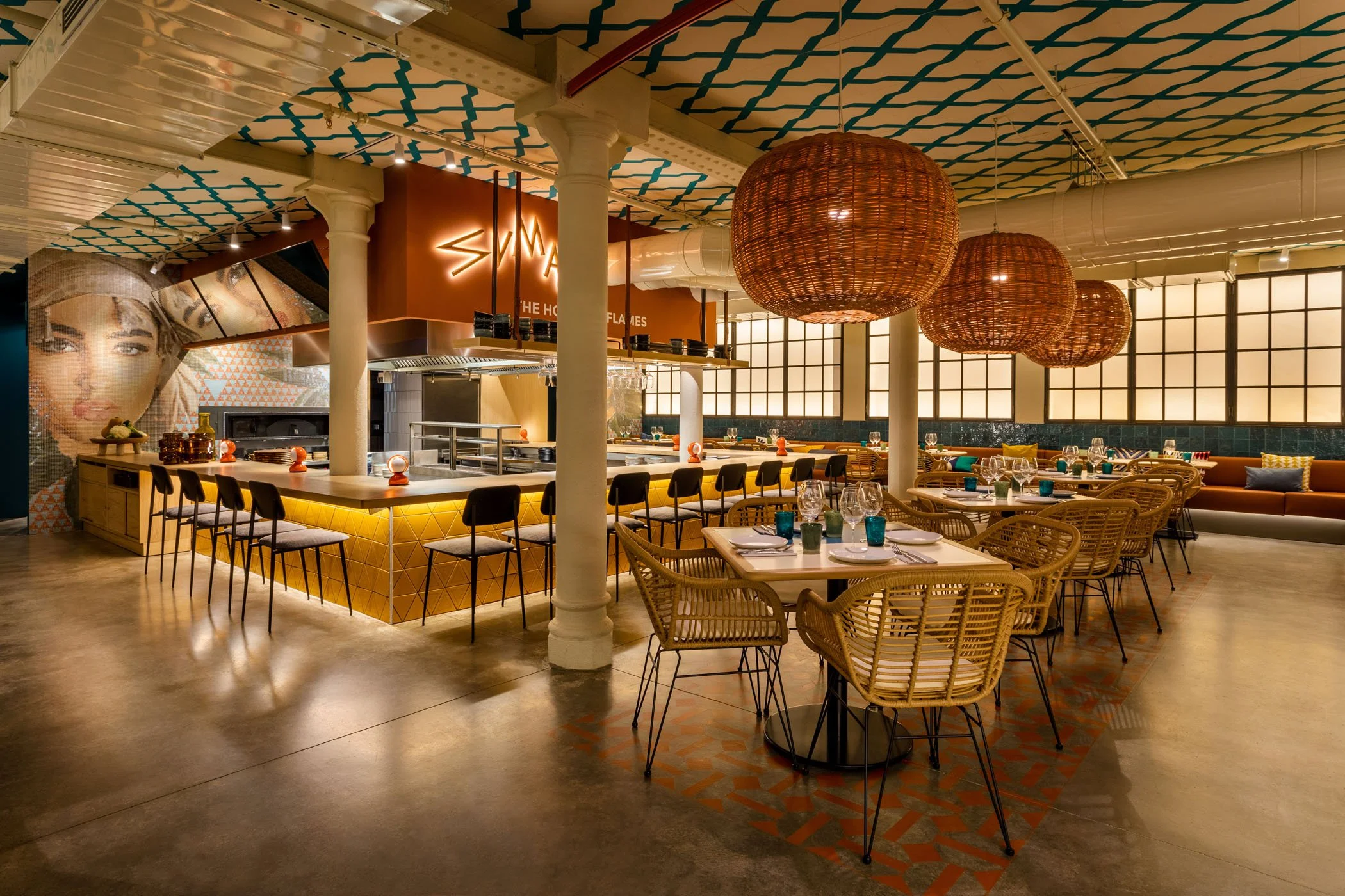 Interior de un restaurante con iluminación cálida, mesas y sillas de mimbre y vidrio, lámparas grandes de fibras naturales y una barra con asientos, decorado con un mural y ventanas con persianas de papel, creando un ambiente acogedor y moderno.