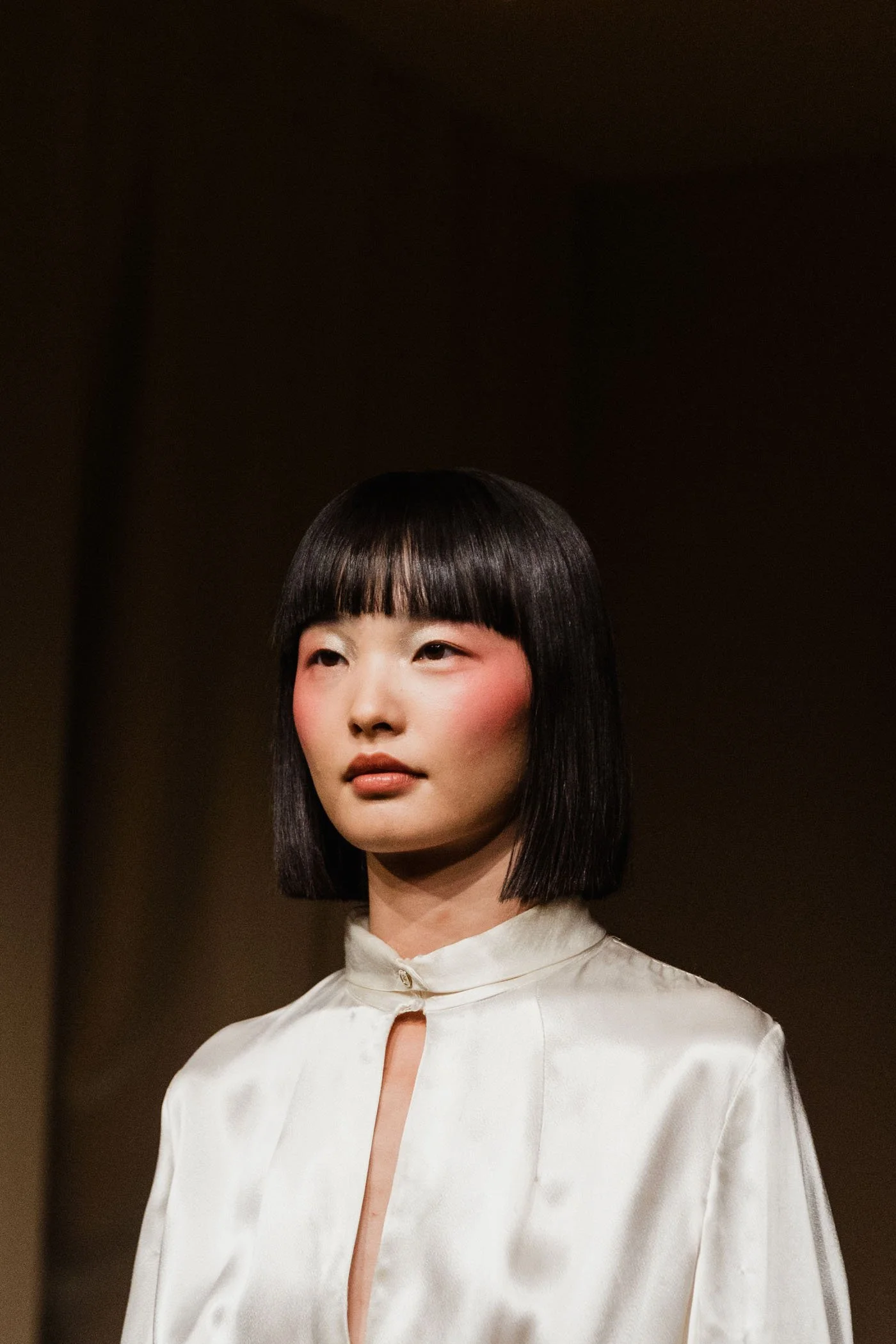 Retrato de una mujer con cabello recto y oscuro, usando blusa blanca con cuello y abertura en el centro, en un fondo oscuro.