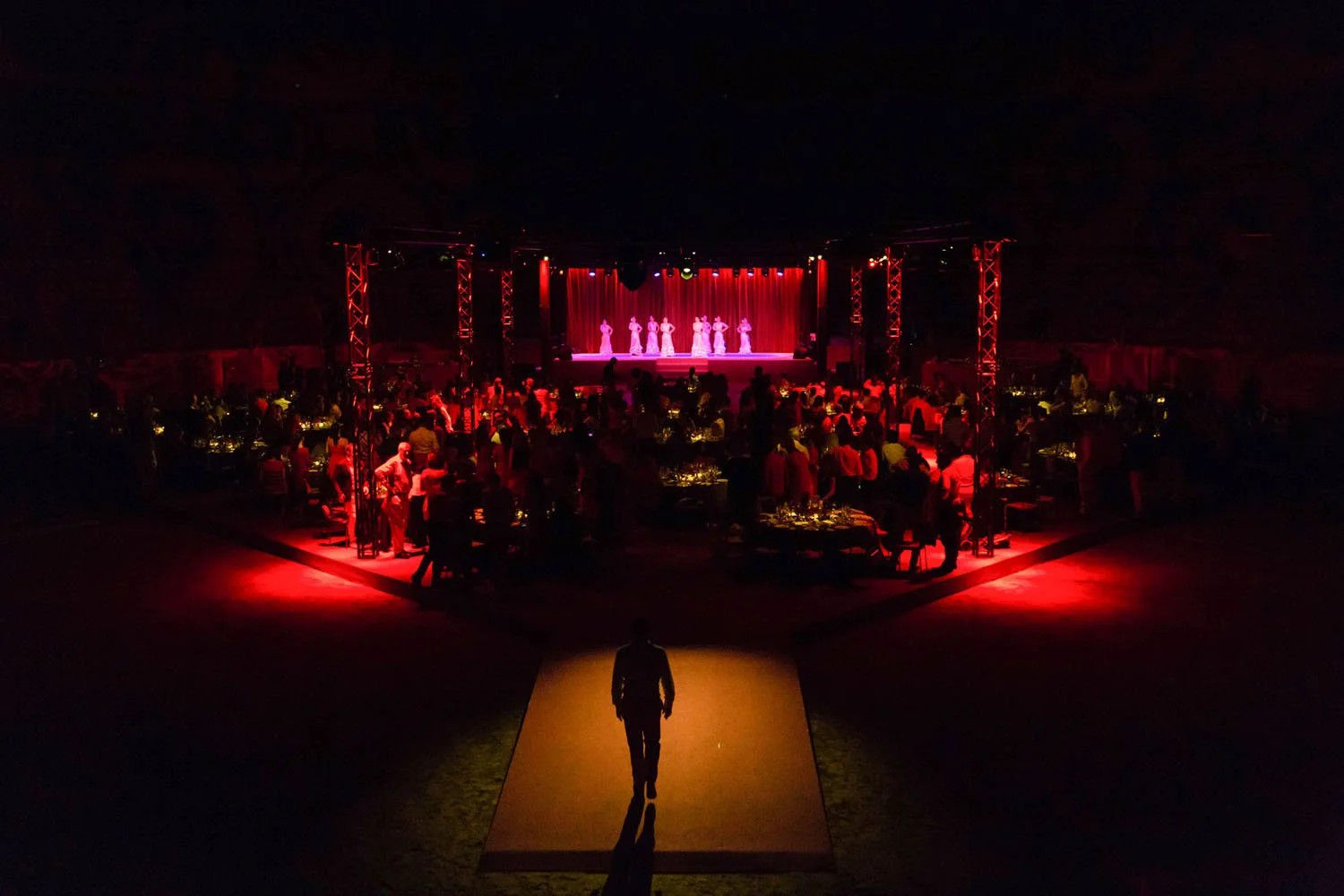 Escenario con bailarines en una presentación, público en mesas, iluminación roja, en un espacio oscuro.