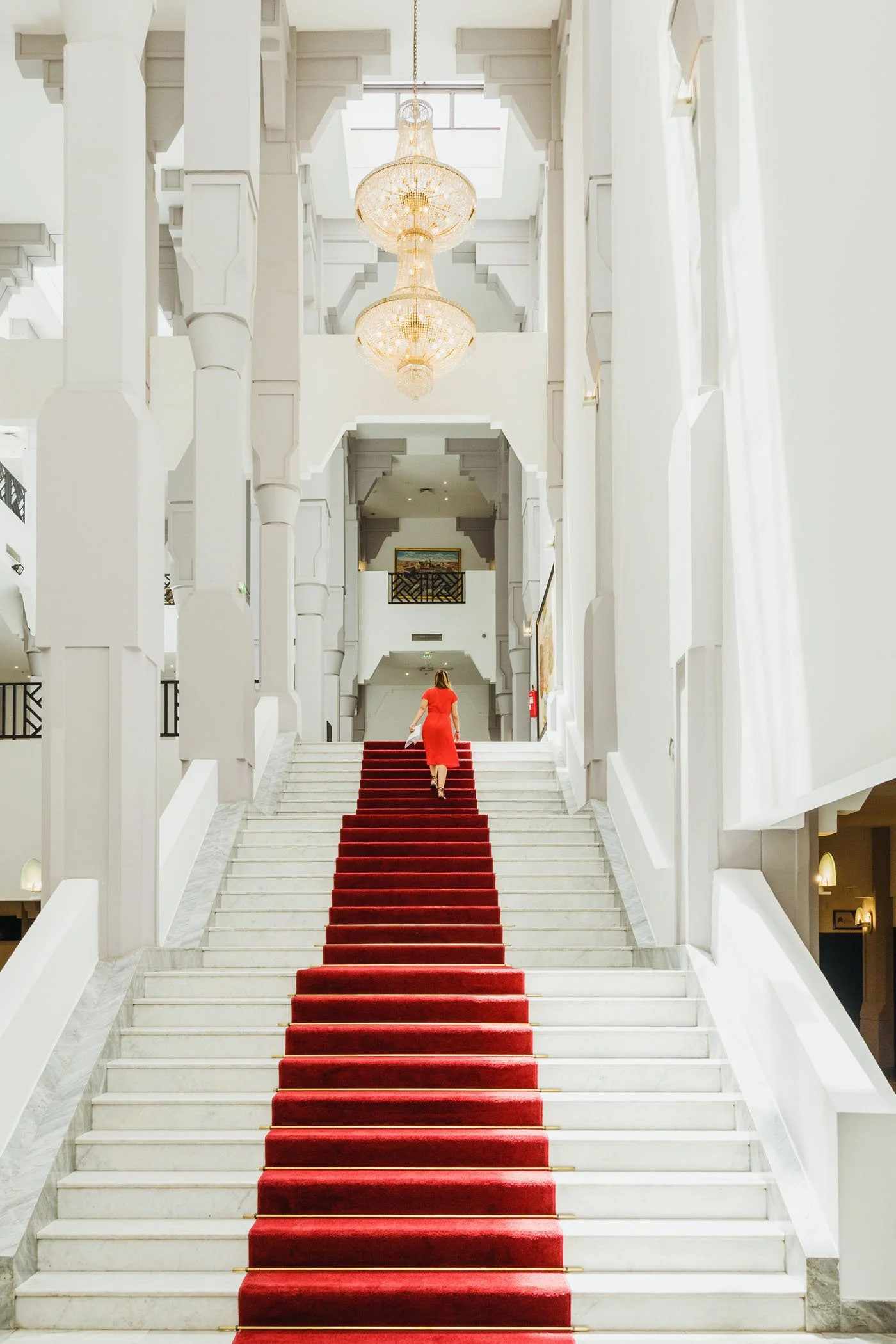 Escalera con alfombra roja en un vestíbulo elegante con paredes blancas y candelabros de techo.