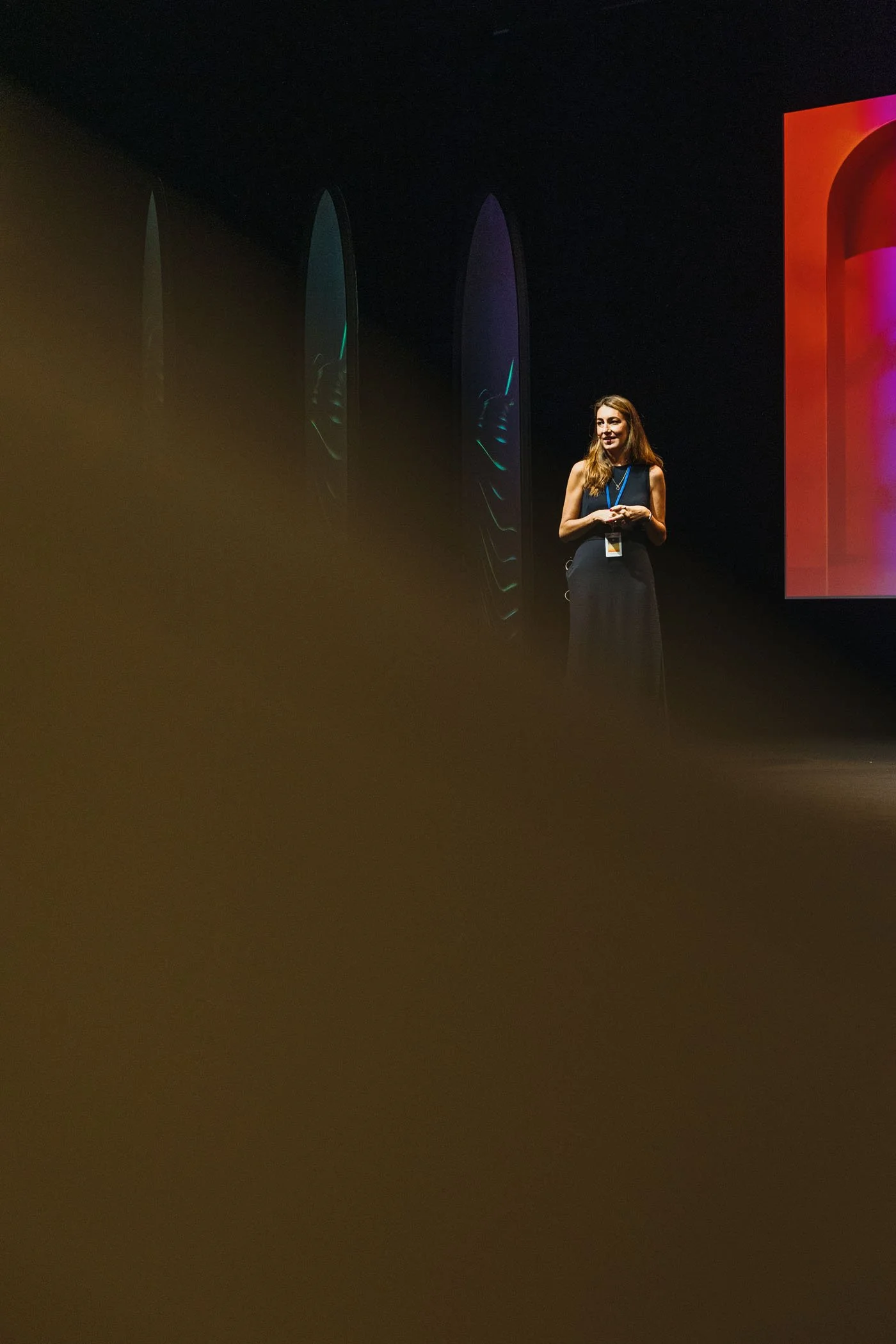 Mujer hablando en una conferencia en un escenario oscuro con una pantalla grande a la derecha y siete formas al fondo.