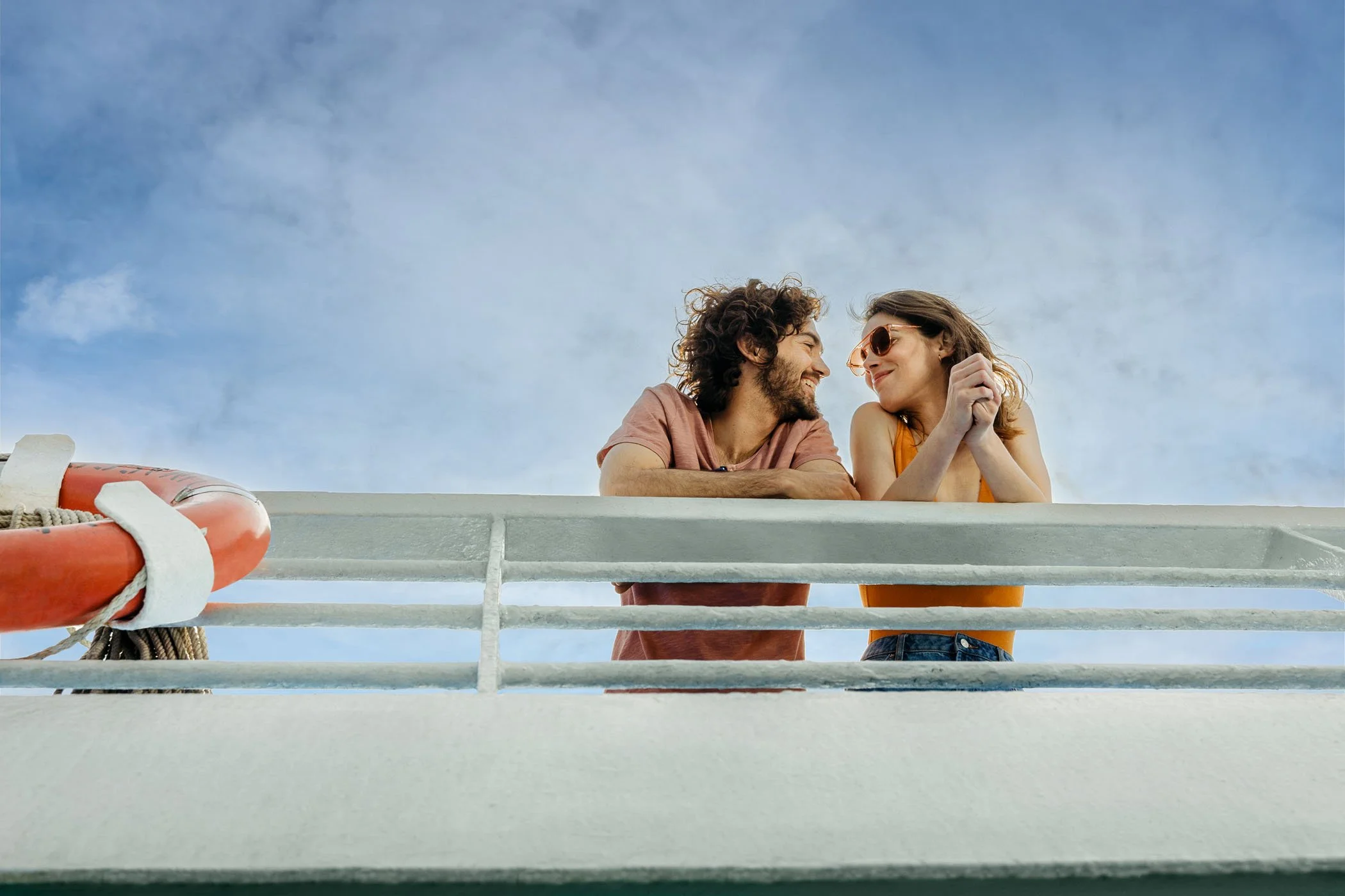 Una pareja sonriendo y mirándose con cariño desde un balcón, mientras el cielo azul con algunas nubes es de fondo.