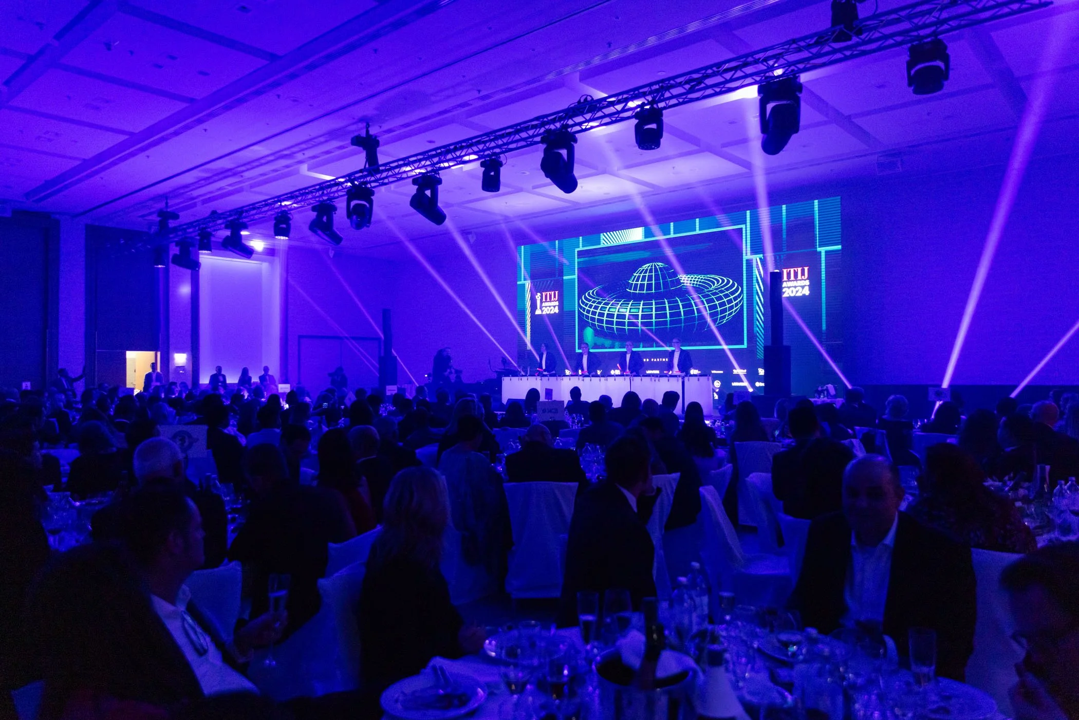 Cena de un evento con muchas personas sentadas en mesas, en un escenario con pantallas que muestran gráficos y el texto 'ITLU Awards 2024', iluminado con luces moradas y azules.