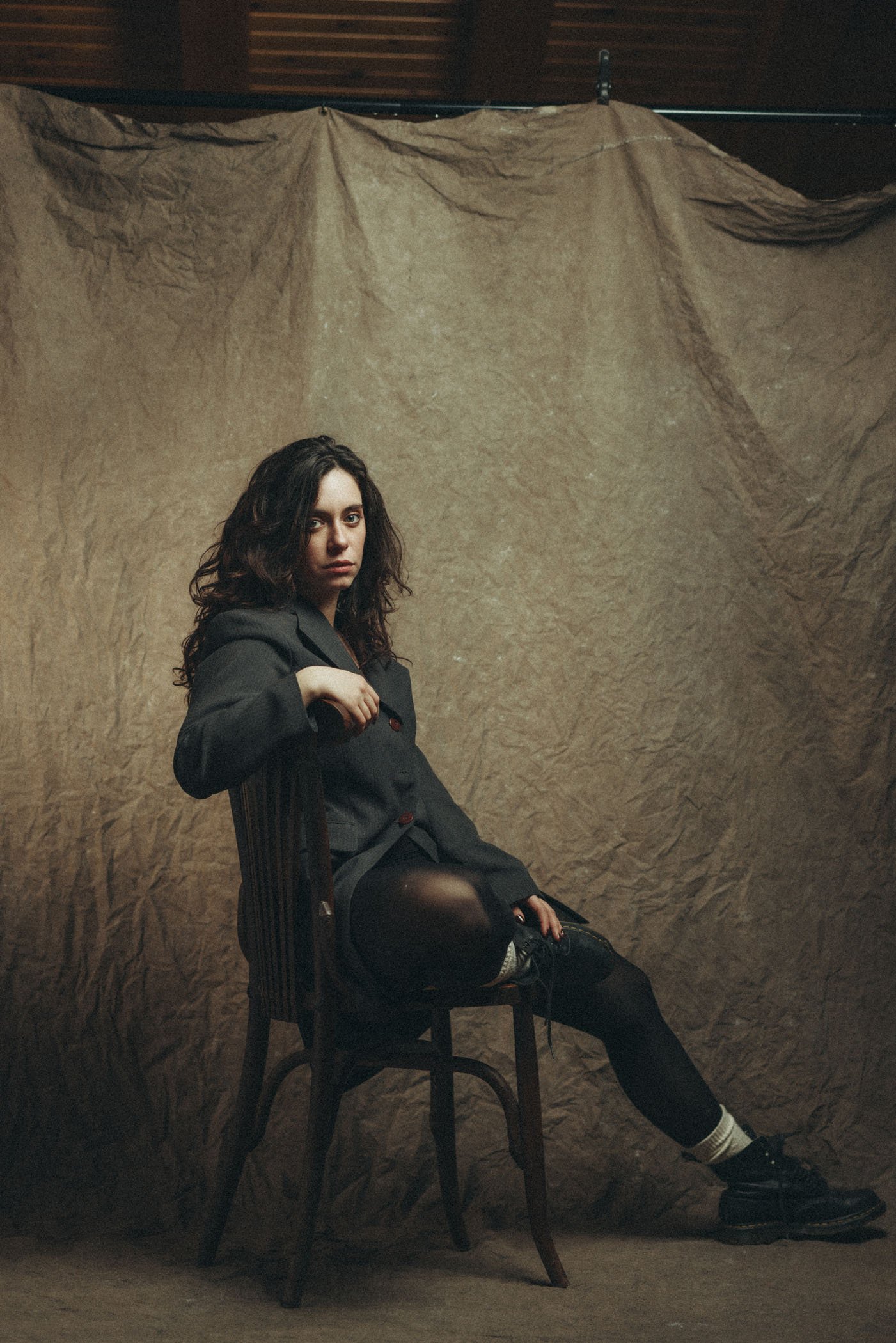 Joven mujer con cabello rizado oscuro, vestida con abrigo gris y pantalones negros, sentada en una silla de madera en un fondo de tela arrugada de color marrón.