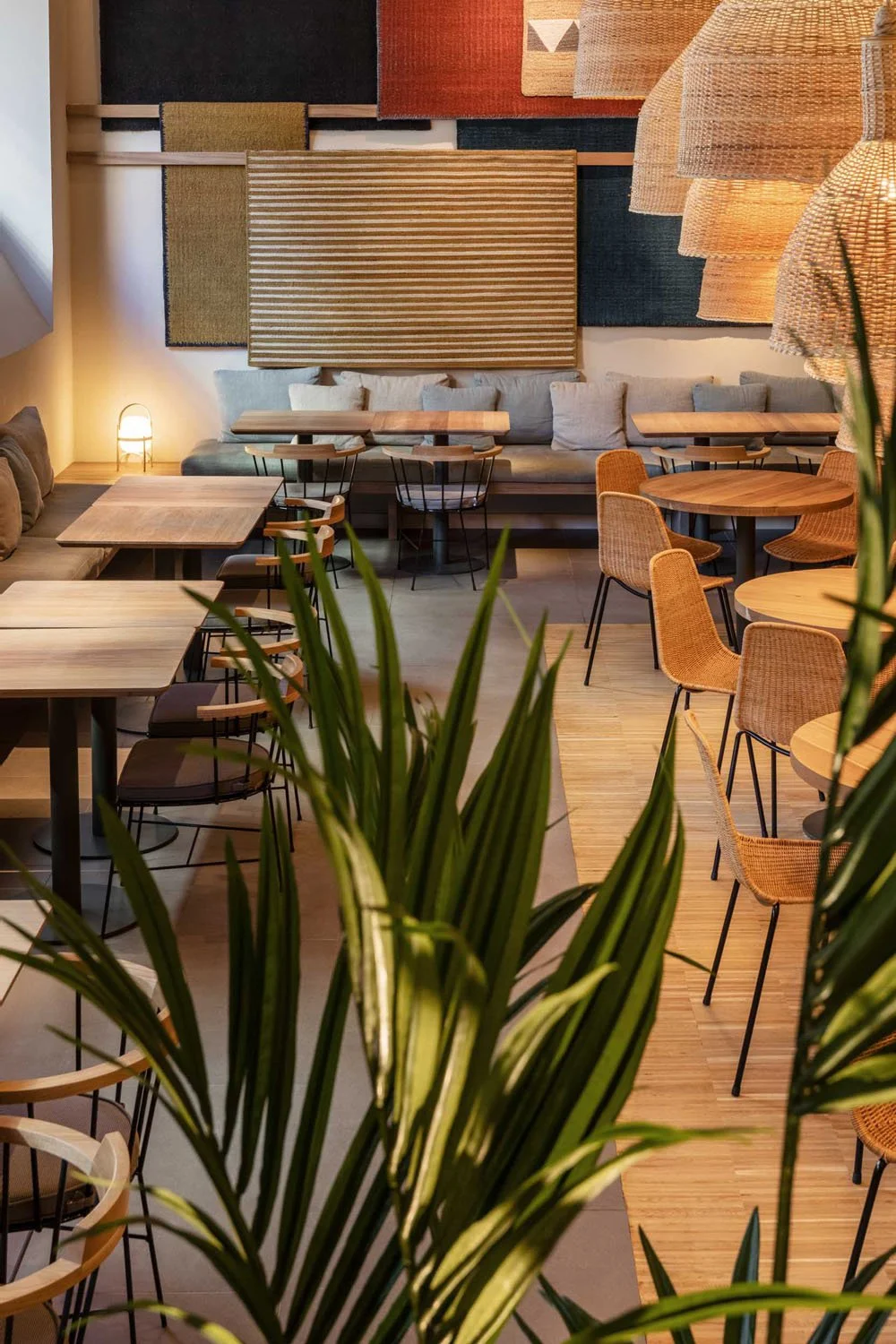 Interior de un café con mesas de madera, sillas de mimbre y una pared decorada con tapices coloridos, con plantas en primer plano.