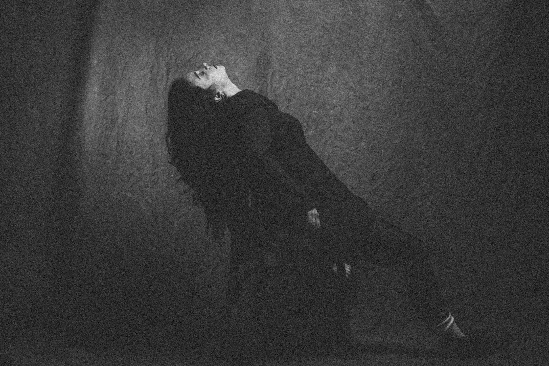 Mujer con cabello oscuro largo, vestida de negro, acostada contra una pared de textura áspera en un ambiente oscuro.