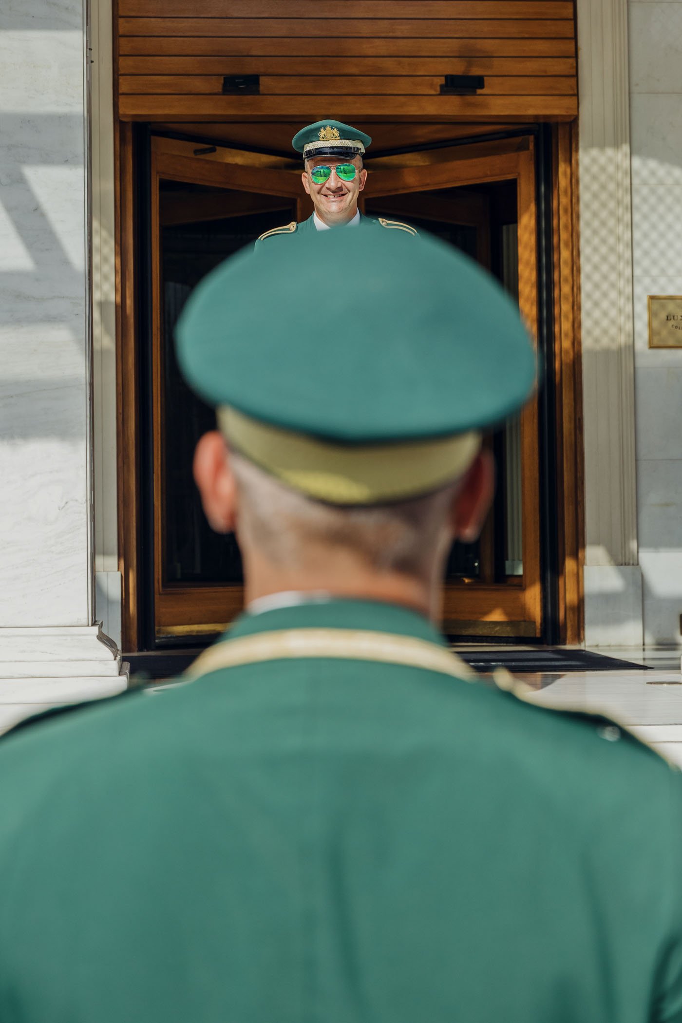 Un oficial con uniforme verde está de espaldas, frente a una persona en uniforme similar que sonríe desde la entrada de un edificio.